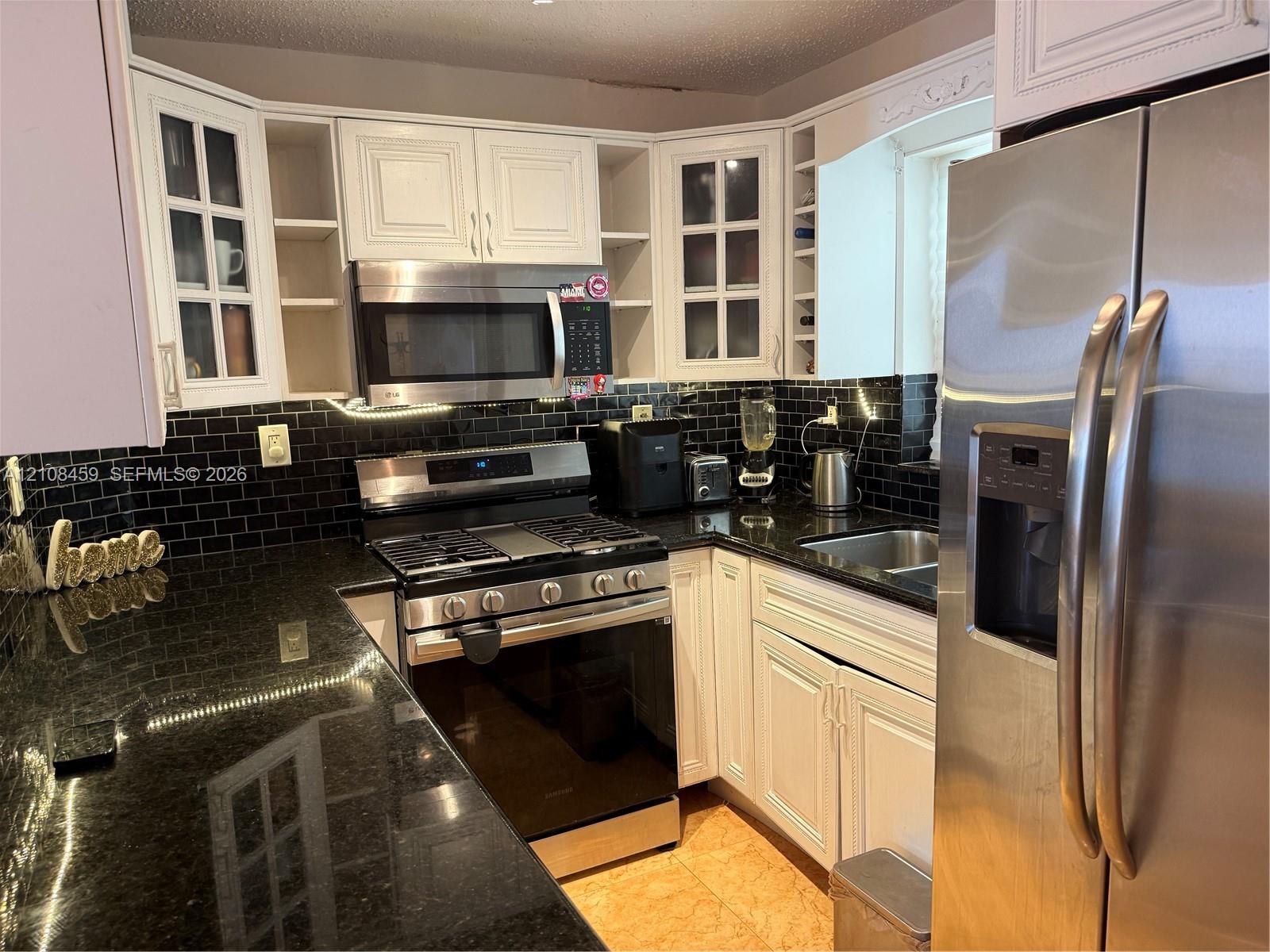 Kitchen featuring white cabinetry with glass-panel display doors, stone countertops, black subway tile backsplash, and stainless steel appliances including a gas range and built-in microwave Kitchen featuring white cabinetry with glass-panel display doors, stone countertops, black subway tile backsplash, and stainless steel appliances including a gas range and built-in microwave