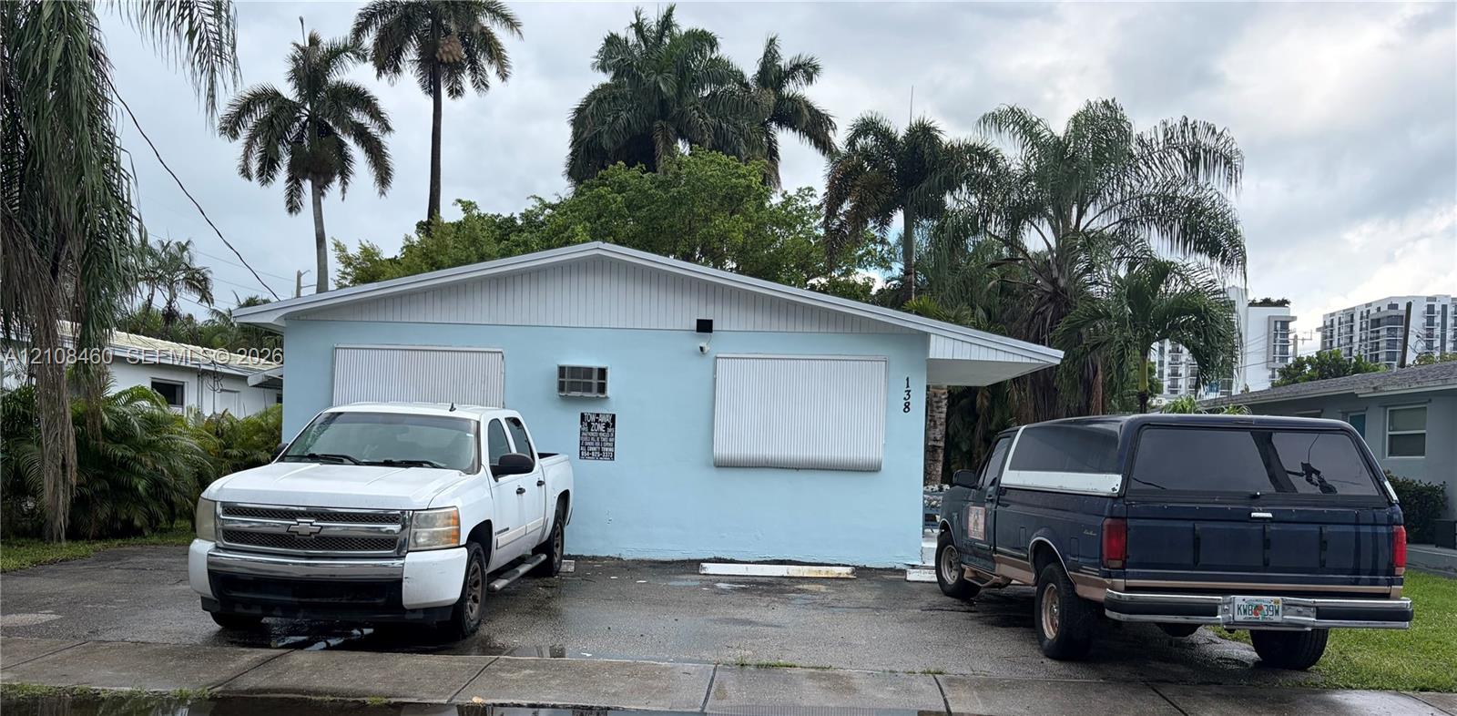 Single-story exterior featuring a light blue facade, a paved parking area, and metal hurricane shutters