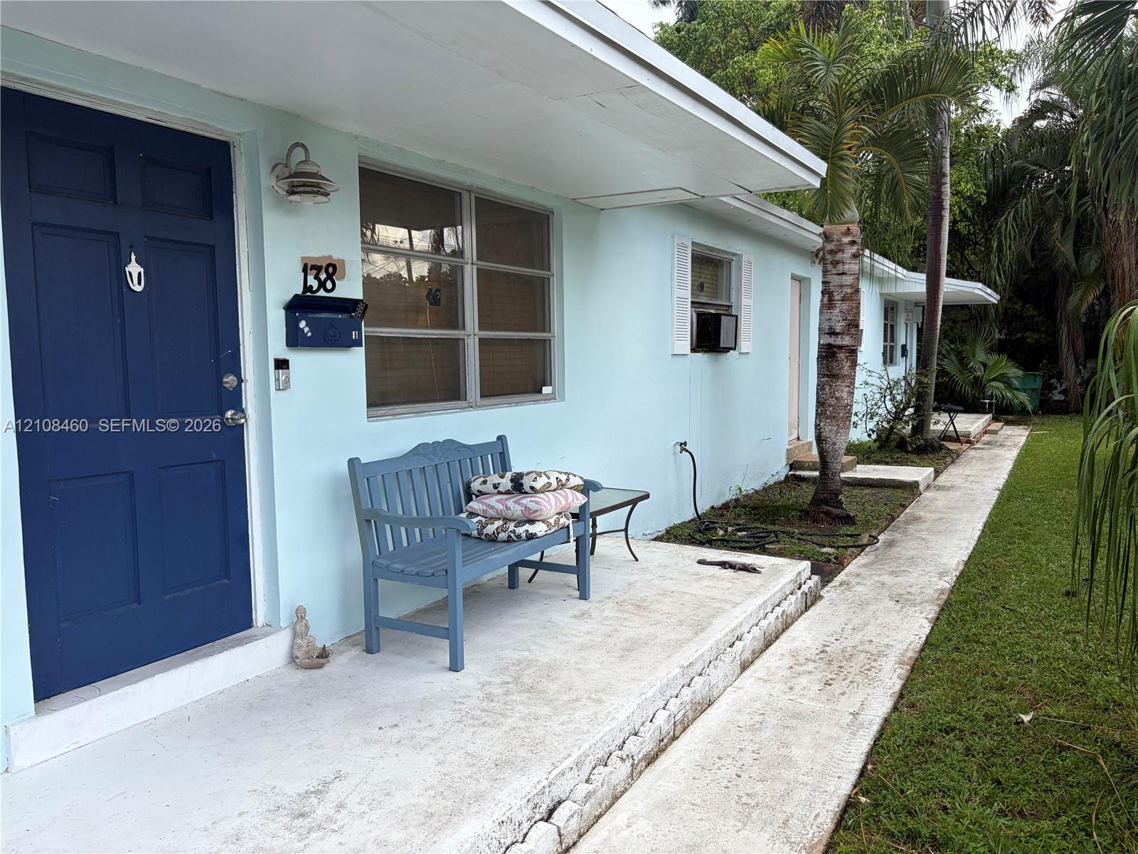 Exterior facade featuring light-toned stucco, a solid blue entry door, an overhanging roof eave, and a concrete pathway leading to a front porch Exterior facade featuring light-toned stucco, a solid blue entry door, an overhanging roof eave, and a concrete pathway leading to a front porch