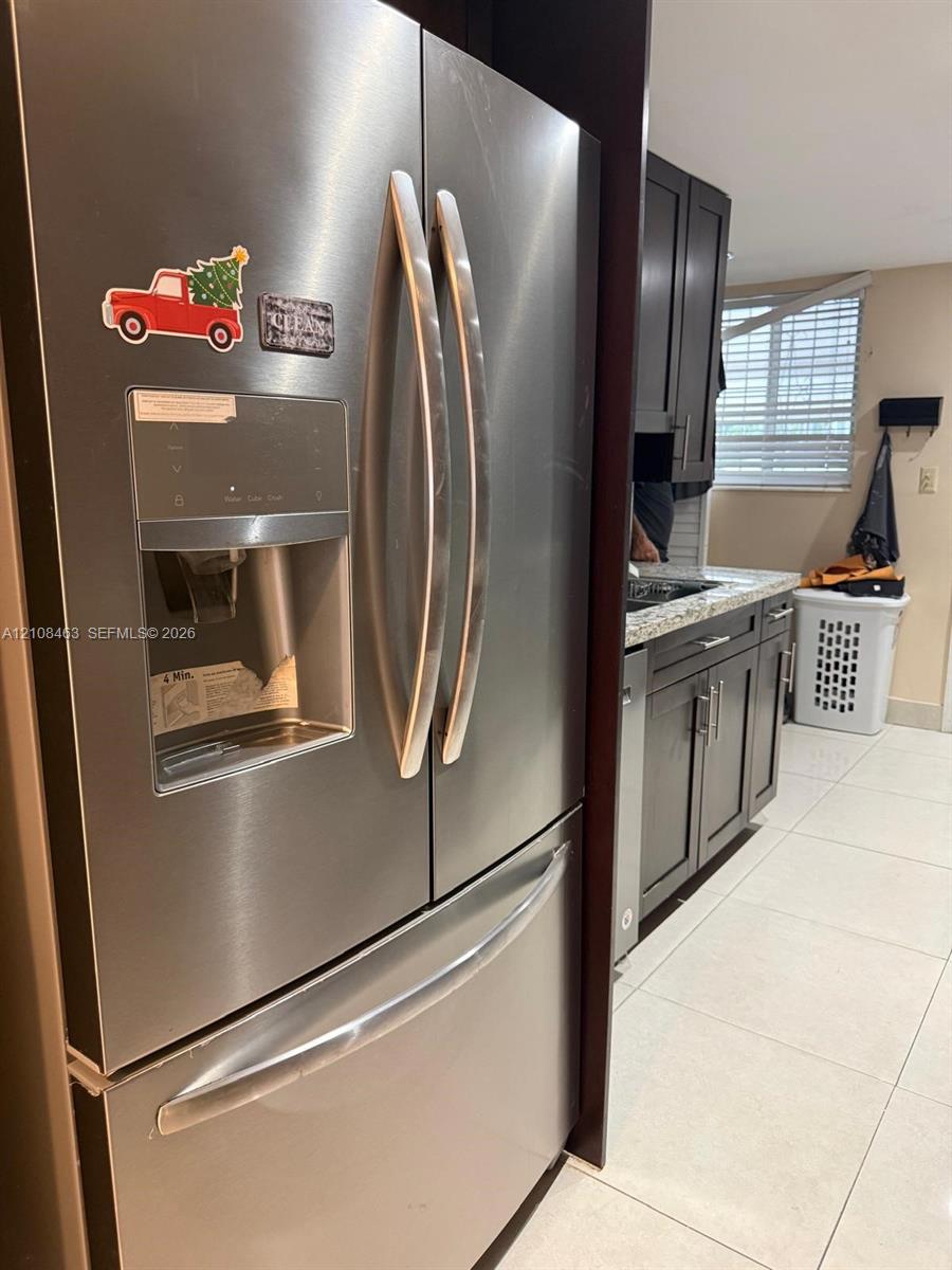 Stainless steel French door refrigerator with exterior water and ice dispenser, dark-toned cabinetry, light-toned stone-finish countertops, and tile flooring Stainless steel French door refrigerator with exterior water and ice dispenser, dark-toned cabinetry, light-toned stone-finish countertops, and tile flooring