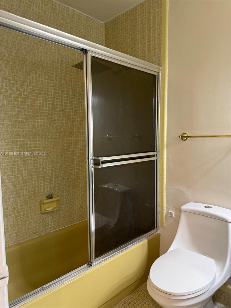 Bathroom featuring a porcelain toilet, a bathtub enclosed by sliding glass doors with metallic trim, grid-patterned wall tile, and an integrated soap dish