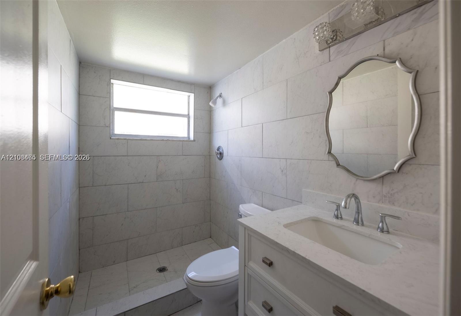 Bathroom featuring stone-finish wall tiling, a vanity with a stone-composite countertop, an integrated sink, polished chrome hardware, and a wall-mounted decorative mirror