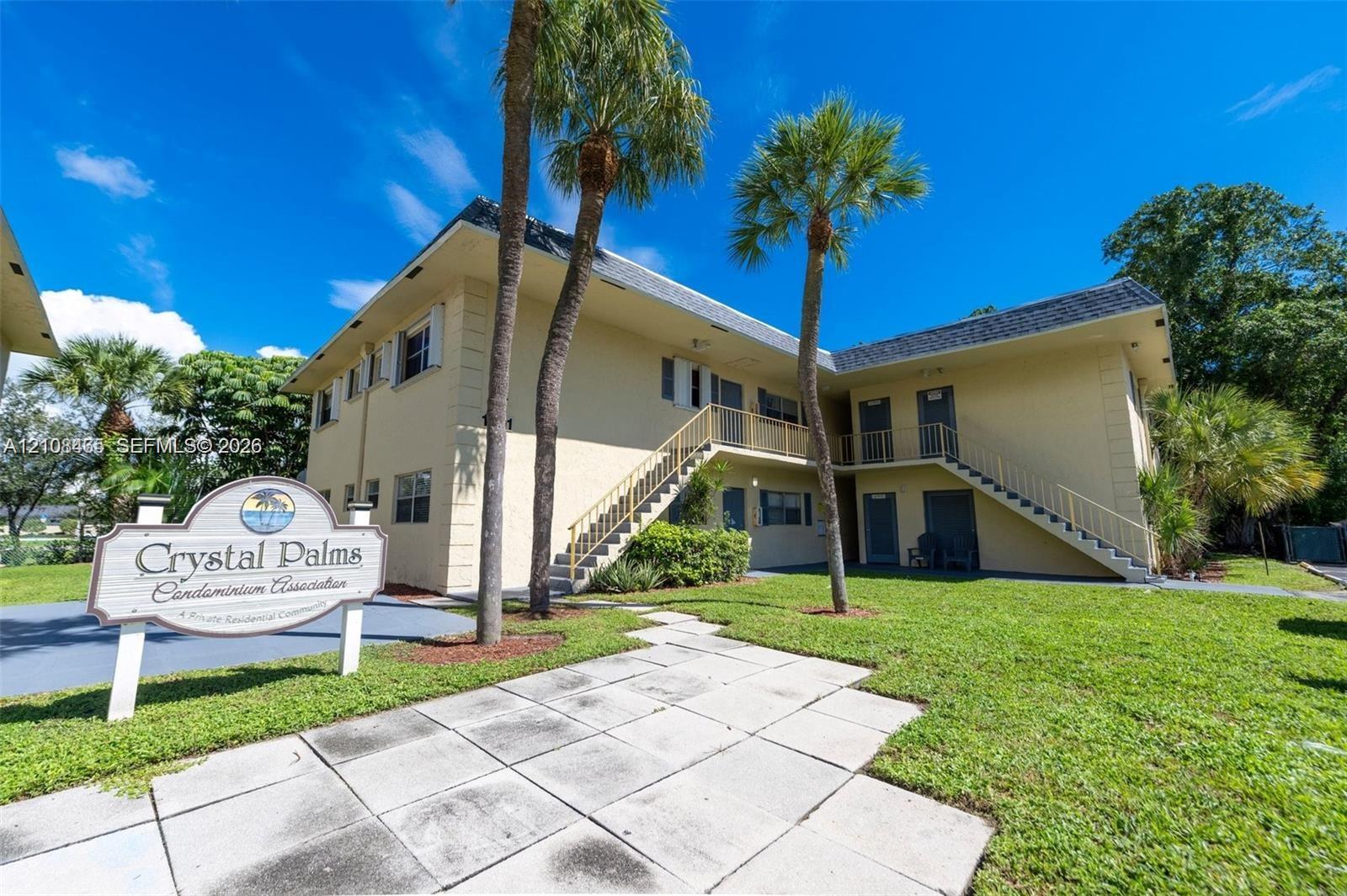 Two-story exterior featuring a light-toned facade, metal staircase railings, manicured lawn, and mature palm trees Two-story exterior featuring a light-toned facade, metal staircase railings, manicured lawn, and mature palm trees