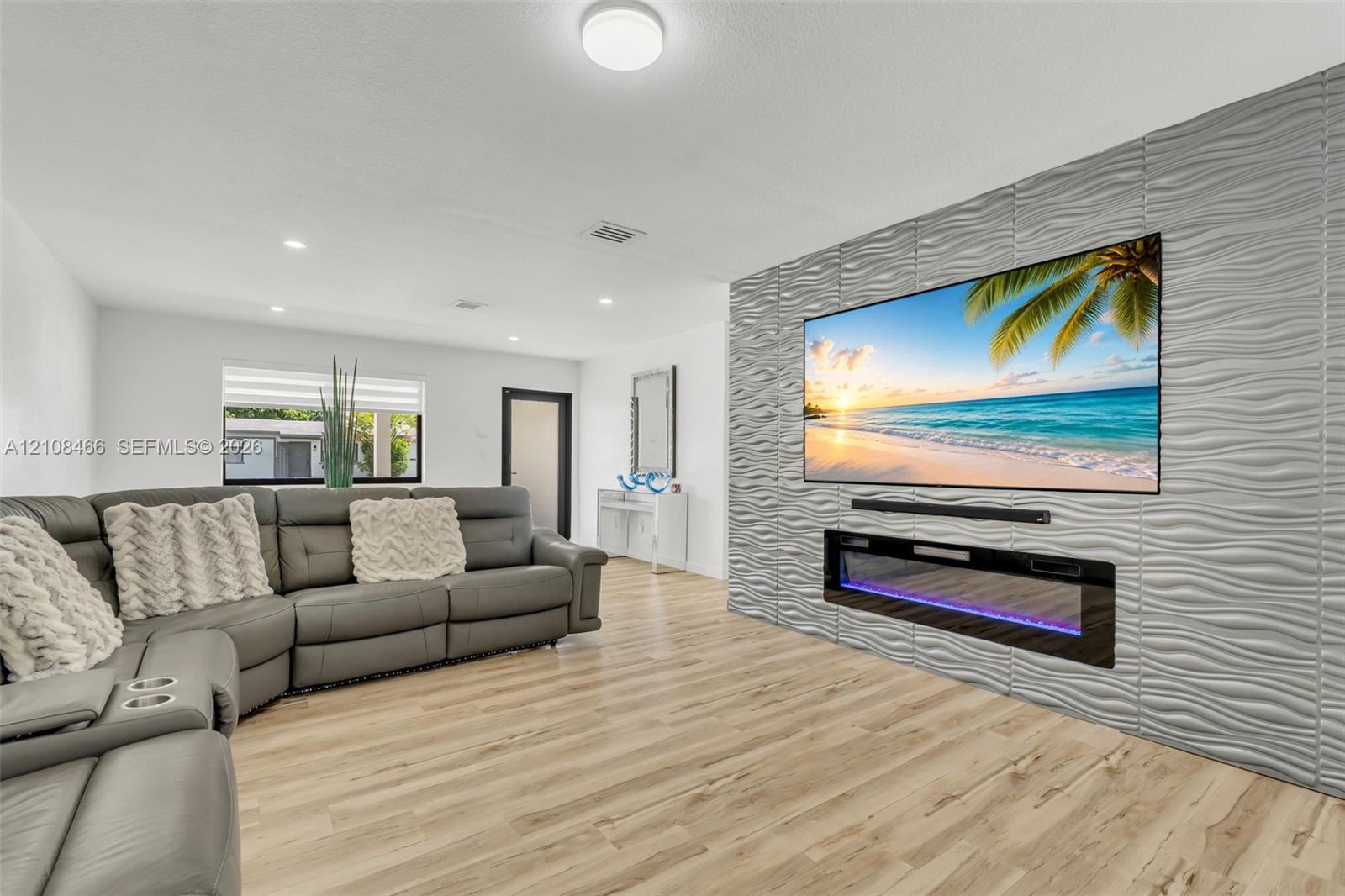 Living area featuring light-toned wood-finish flooring, a textured 3D accent wall, and a built-in linear electric fireplace Living area featuring light-toned wood-finish flooring, a textured 3D accent wall, and a built-in linear electric fireplace
