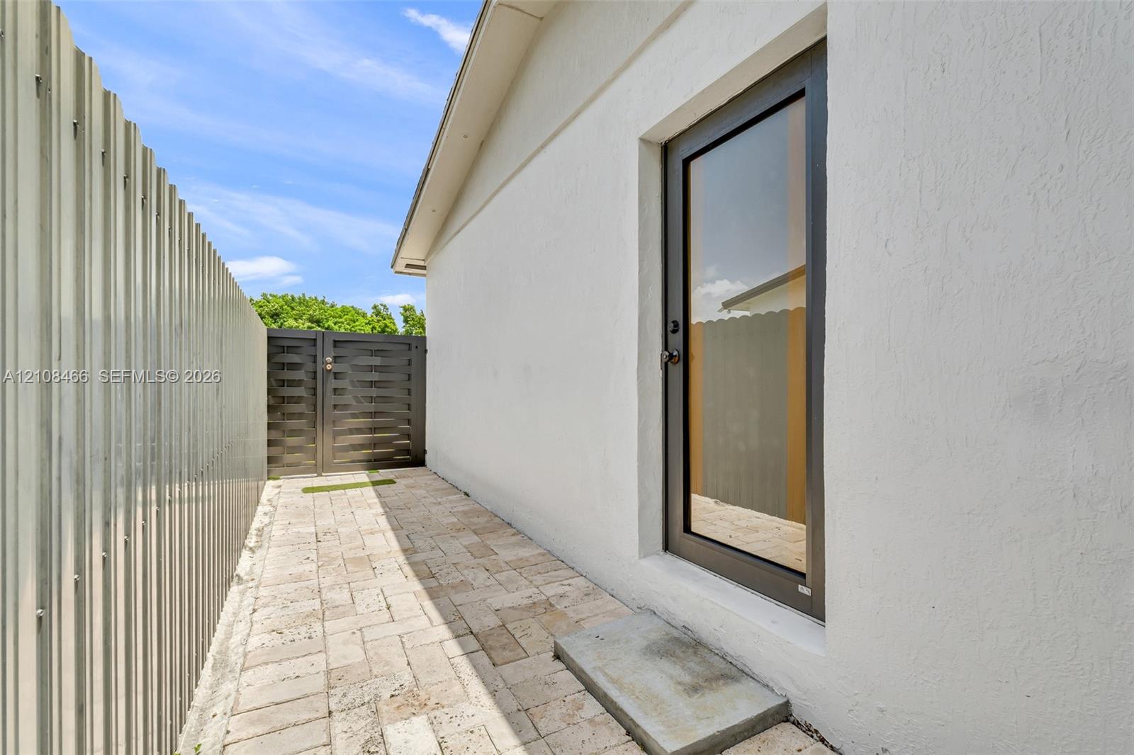 Exterior side passage featuring light-toned paver hardscaping, a full-lite glass door with a metallic frame, a corrugated metal privacy fence, and a horizontal-slat metal gate