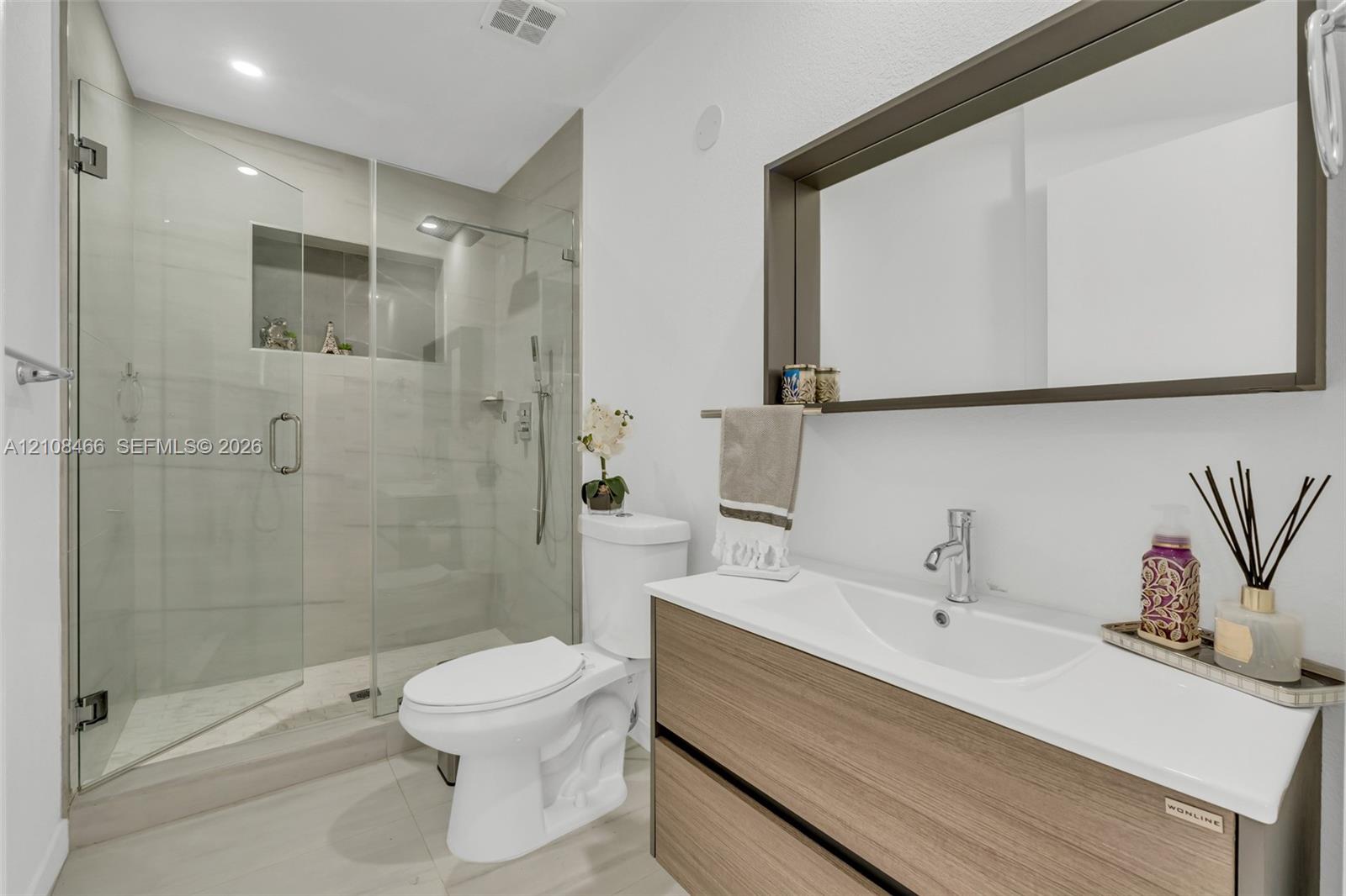 Floating wood-finish vanity with an integrated sink, frameless glass walk-in shower featuring a tiled inset niche, and a wall-mounted framed mirror