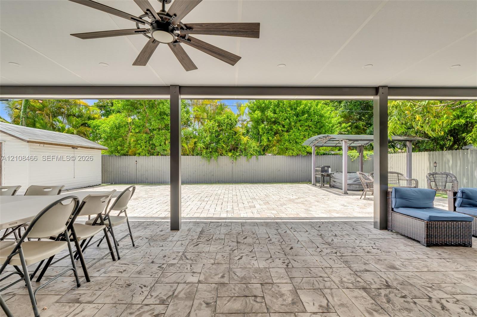 Covered patio featuring a stamped concrete floor, recessed lighting, and a multi-blade ceiling fan, leading to a brick-paved backyard with a perimeter fence