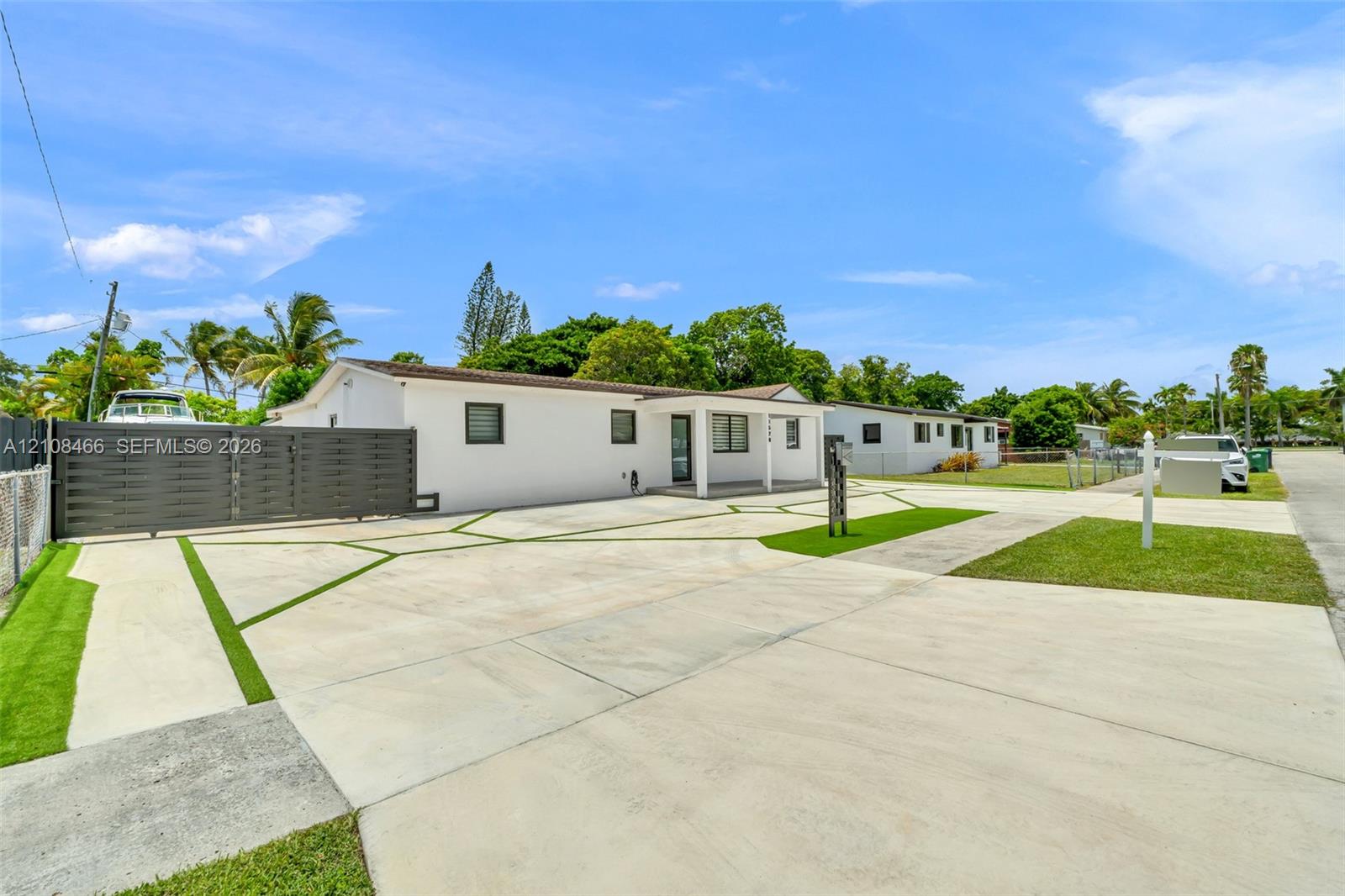 Single-story residence featuring white stucco exterior, expansive concrete driveway with geometric turf accents, horizontal slat privacy fencing, and covered front entryway Single-story residence featuring white stucco exterior, expansive concrete driveway with geometric turf accents, horizontal slat privacy fencing, and covered front entryway