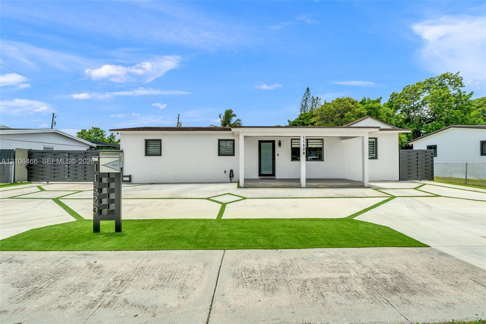 Single-story exterior featuring white stucco siding, a covered front porch, geometric concrete driveway with synthetic grass inlays, and dual metal privacy fencing Single-story exterior featuring white stucco siding, a covered front porch, geometric concrete driveway with synthetic grass inlays, and dual metal privacy fencing