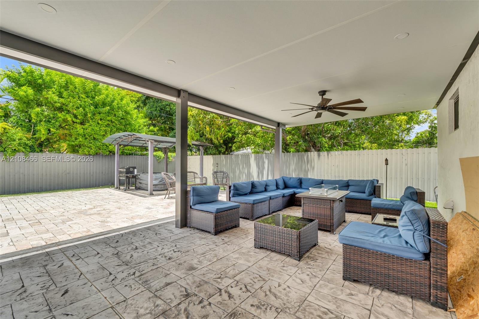 Covered patio featuring stamped concrete flooring, recessed lighting, and a ceiling fan, opening onto a paved outdoor area with a metal-framed structure and privacy fencing
