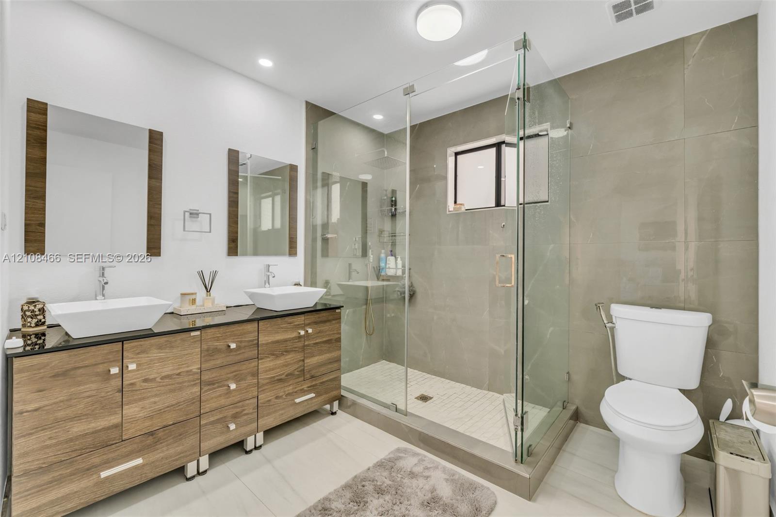 Dual vanity with rectangular vessel sinks, dark stone countertop, wood-finish cabinetry, walk-in glass-enclosed shower featuring full-height tile surround, and recessed lighting