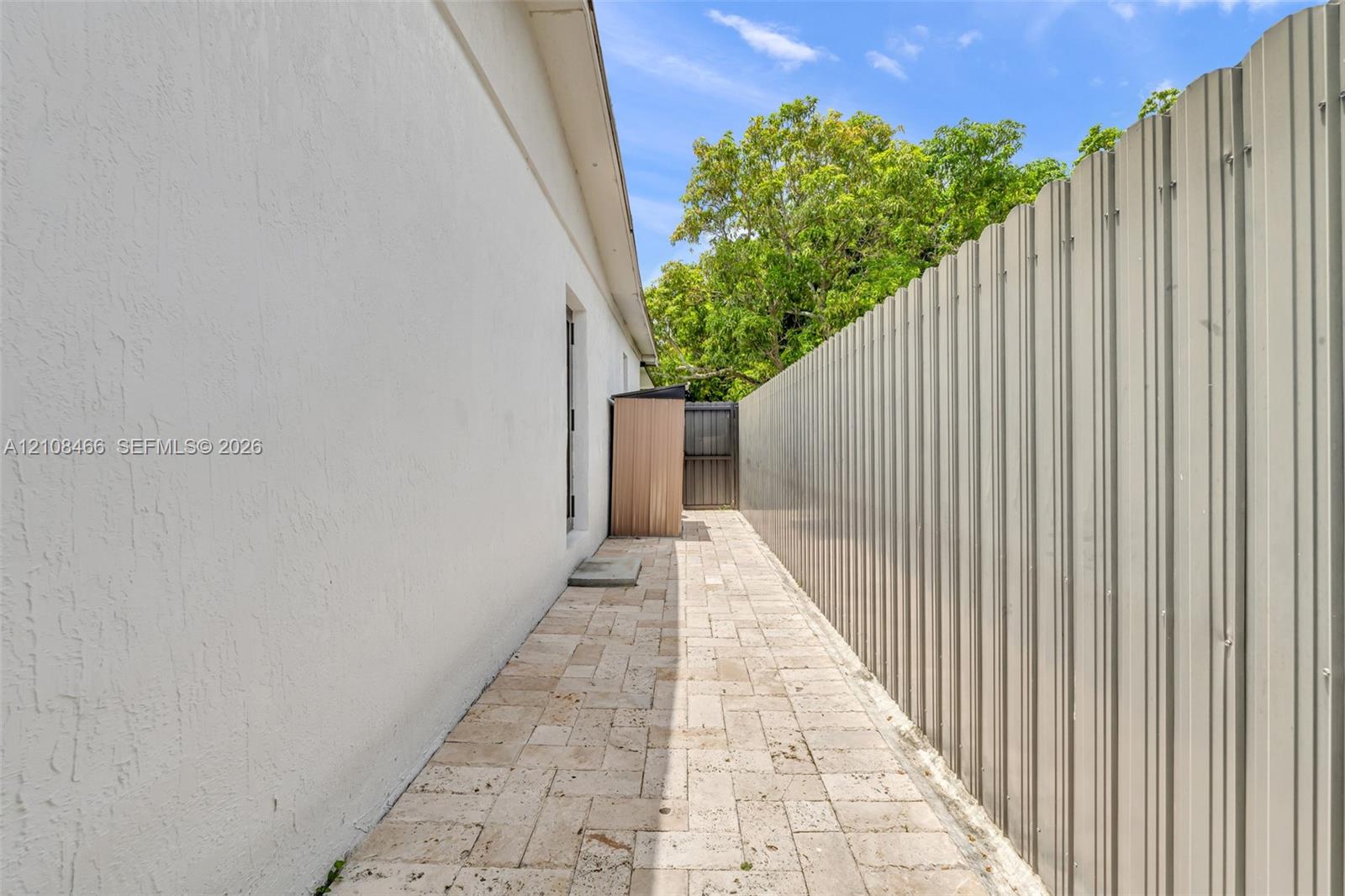Exterior side yard featuring light-colored stone pavers and a corrugated metal privacy fence