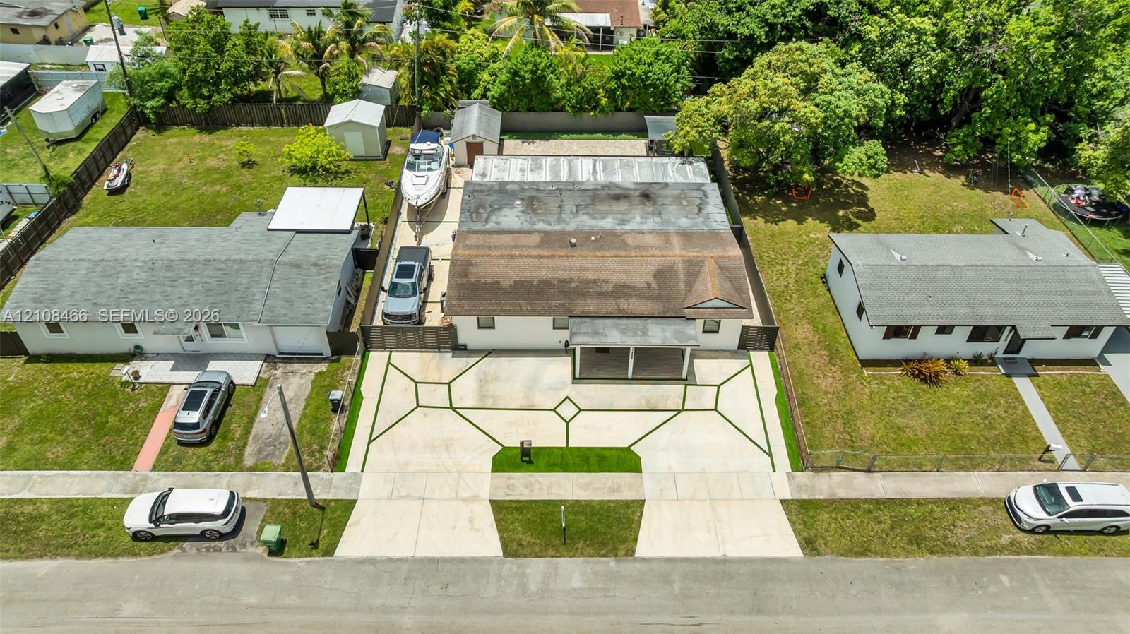Aerial perspective of property featuring an expansive concrete driveway with decorative geometric grass inlays, a covered front porch, and a fenced backyard with detached storage structures