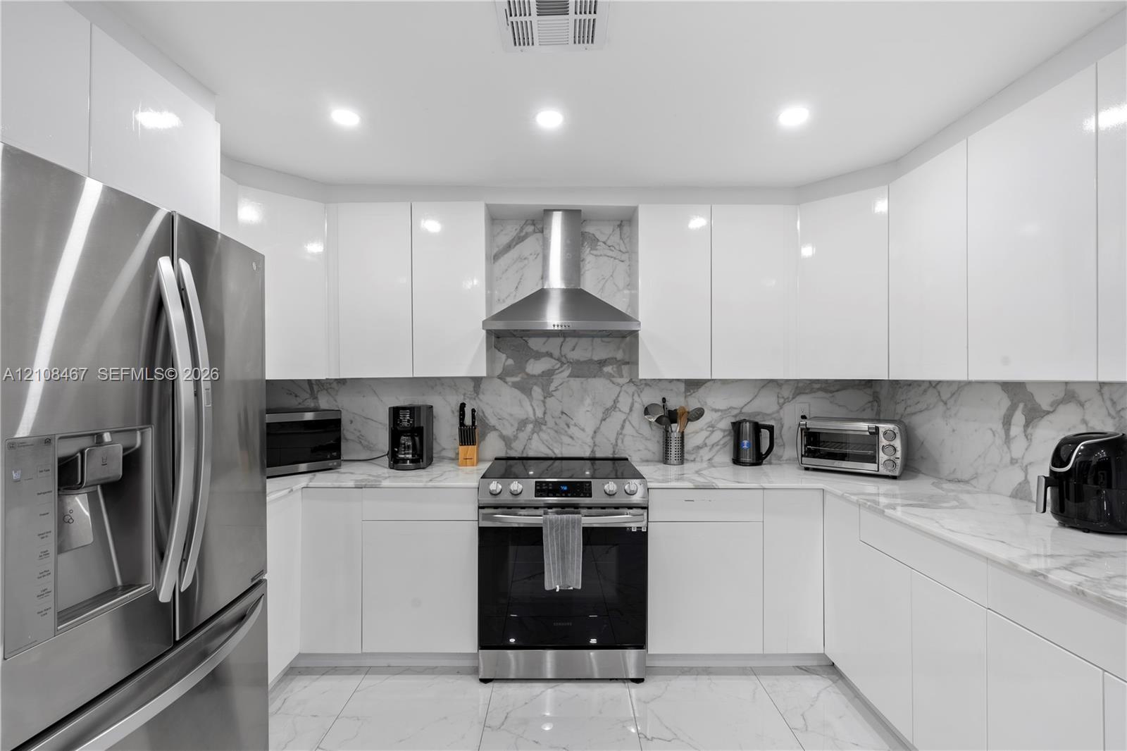 Modern kitchen featuring high-gloss white cabinetry, stone-surface countertops, full-height stone backsplash, stainless steel appliances, and large-format tile flooring