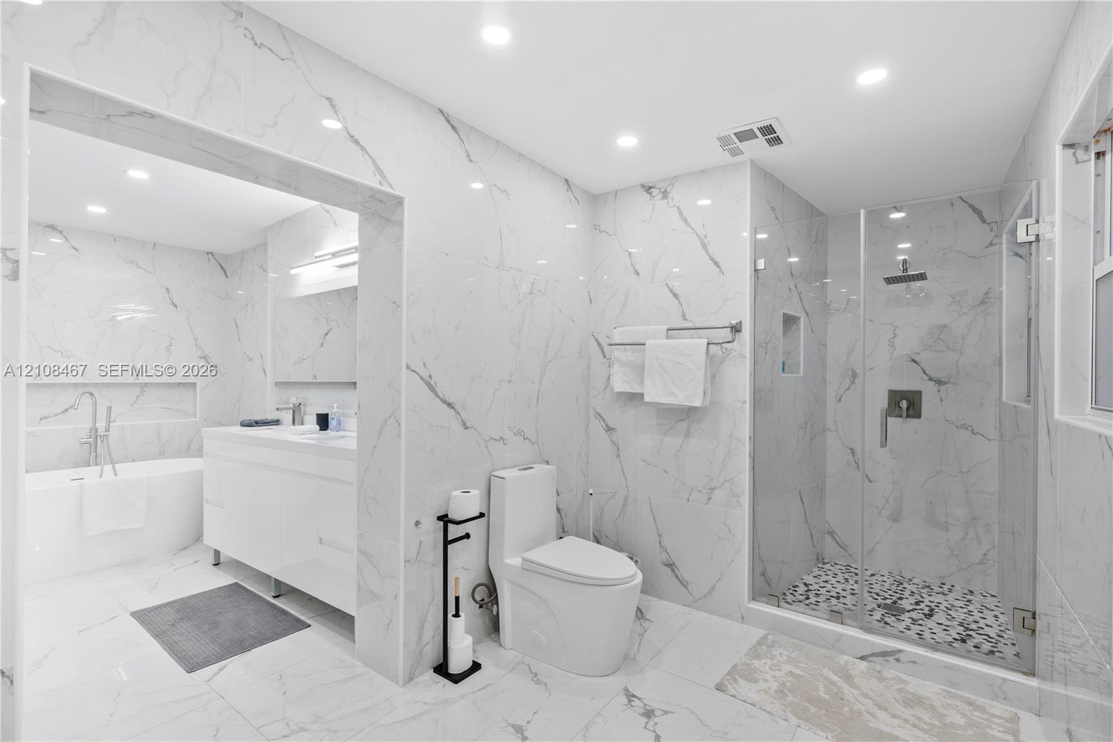 Full-height wall-to-wall stone-finish tile, glass-enclosed shower with pebble-finish flooring, built-in recessed wall niche, freestanding soaking tub, and double-vanity cabinet