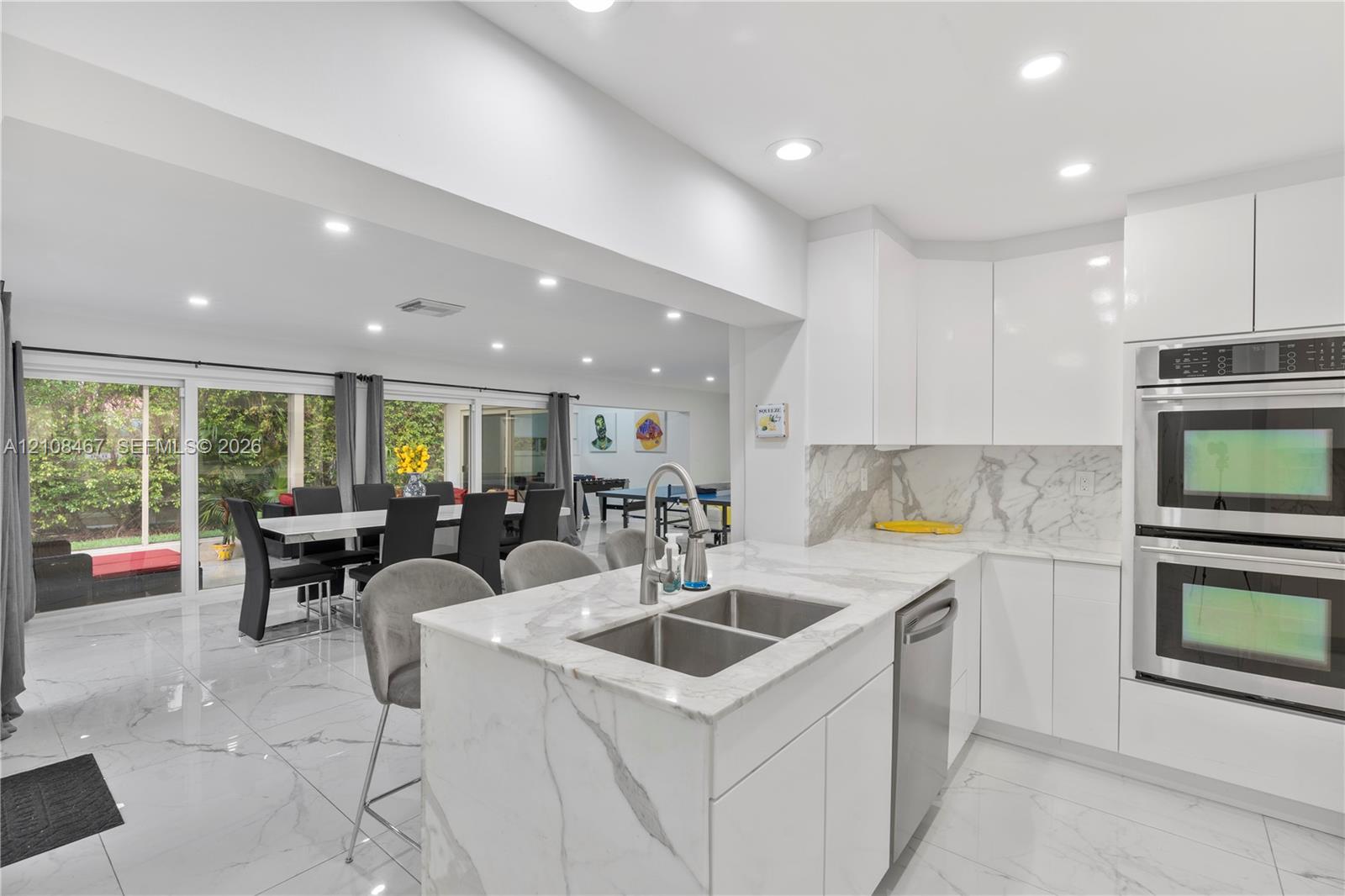 Kitchen featuring white flat-panel cabinetry, stone-patterned countertops, a central island with a double-basin stainless steel sink, built-in double wall ovens, and large-format tile flooring