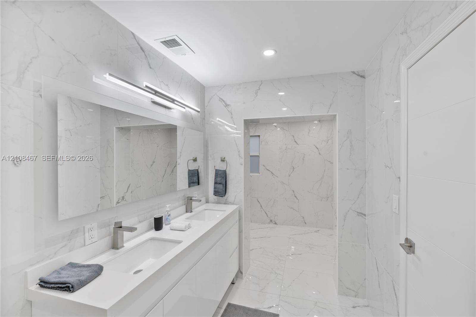Dual-sink vanity with white countertop, full-width mirror, integrated wall-mounted lighting, floor-to-ceiling stone-look wall tiling, and walk-in shower area