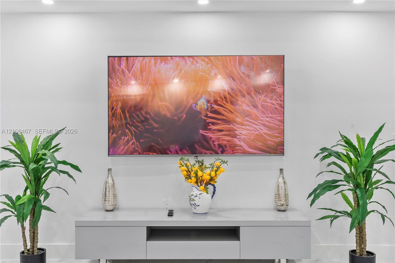 Wall-mounted display screen above a minimalist, light gray media console featuring integrated storage cabinetry and recessed ceiling lighting