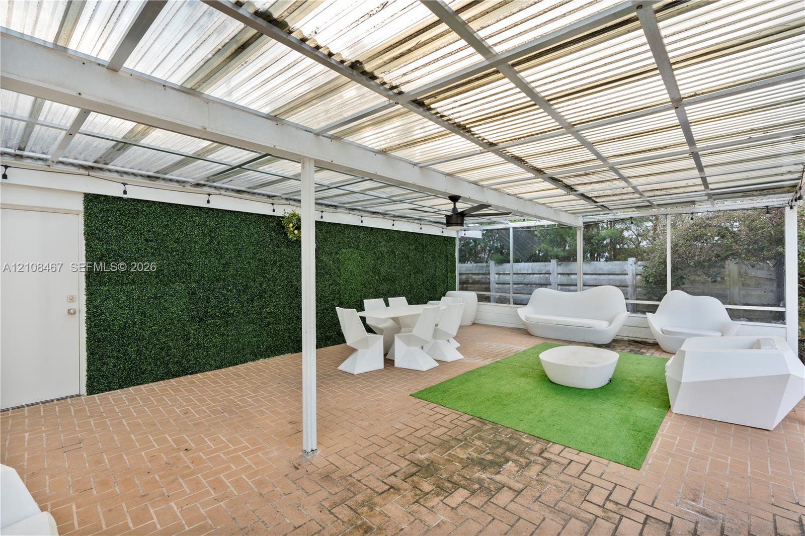 Covered patio featuring a corrugated translucent roof, brick paver flooring, green decorative privacy wall, ceiling fan, and white structural support posts