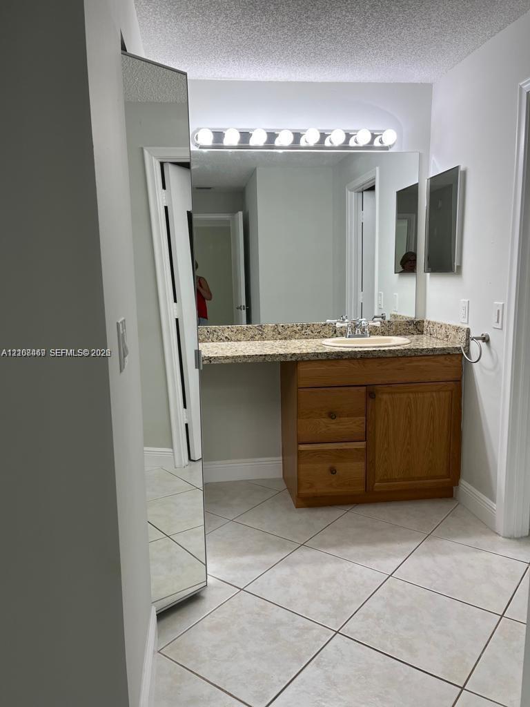 Vanity featuring wooden cabinetry, stone-finish countertop, oval undermount sink, wall-spanning mirror with integrated light fixture, and diagonal tile flooring.