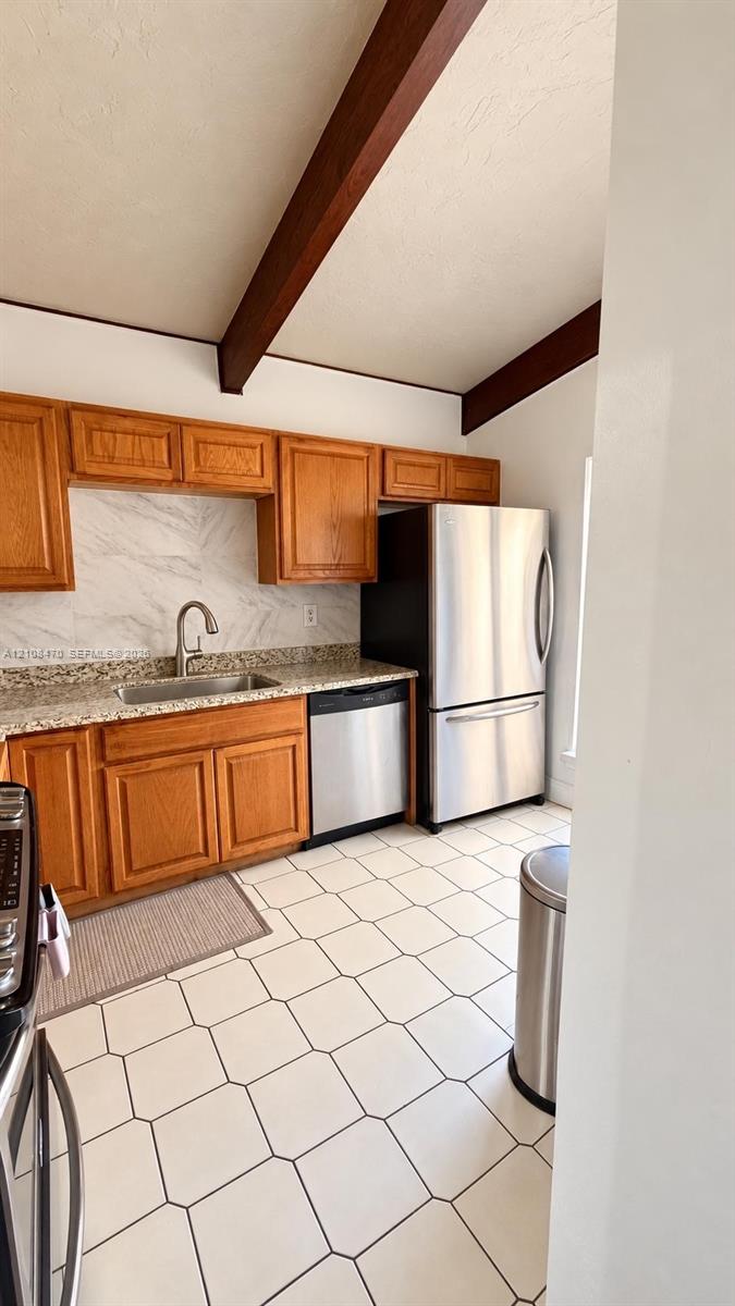 Kitchen featuring wood cabinetry, stone-finish countertops, stainless steel appliances, ceramic tile flooring, and vaulted ceilings with exposed wood beams