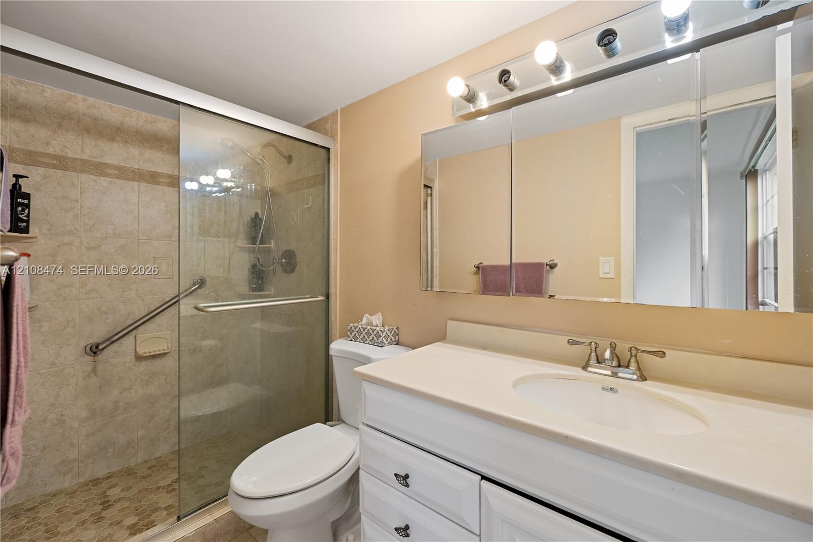 Bathroom featuring a vanity with white cabinetry, a light-toned countertop, an integrated sink, a glass-enclosed shower with tiled surround, and a tri-fold medicine cabinet with attached vanity lighting