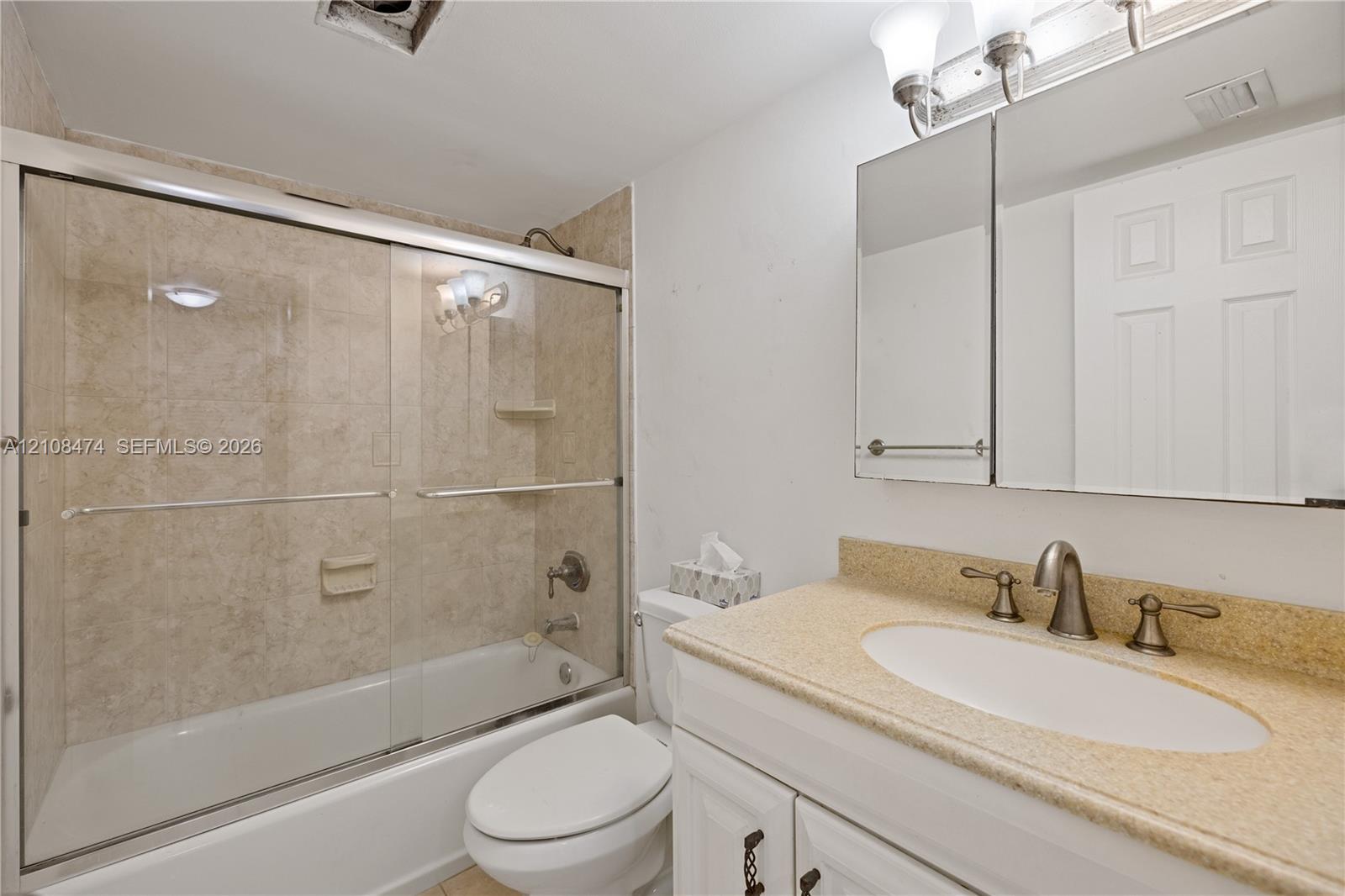 Bathroom featuring a vanity with a solid-surface countertop, an undermount oval sink, metallic finish widespread faucet, mirrored medicine cabinet, and a shower-tub combination with sliding glass doors and tiled surround