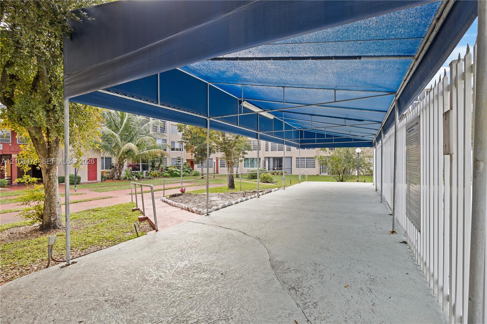 Covered outdoor patio featuring a metal frame structure, blue shade canopy, concrete flooring, and perimeter metal fencing