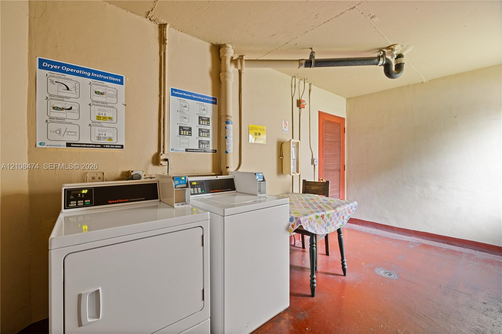 Utility area featuring two industrial-style, top-load laundry appliances, painted concrete flooring, and exposed overhead plumbing fixtures