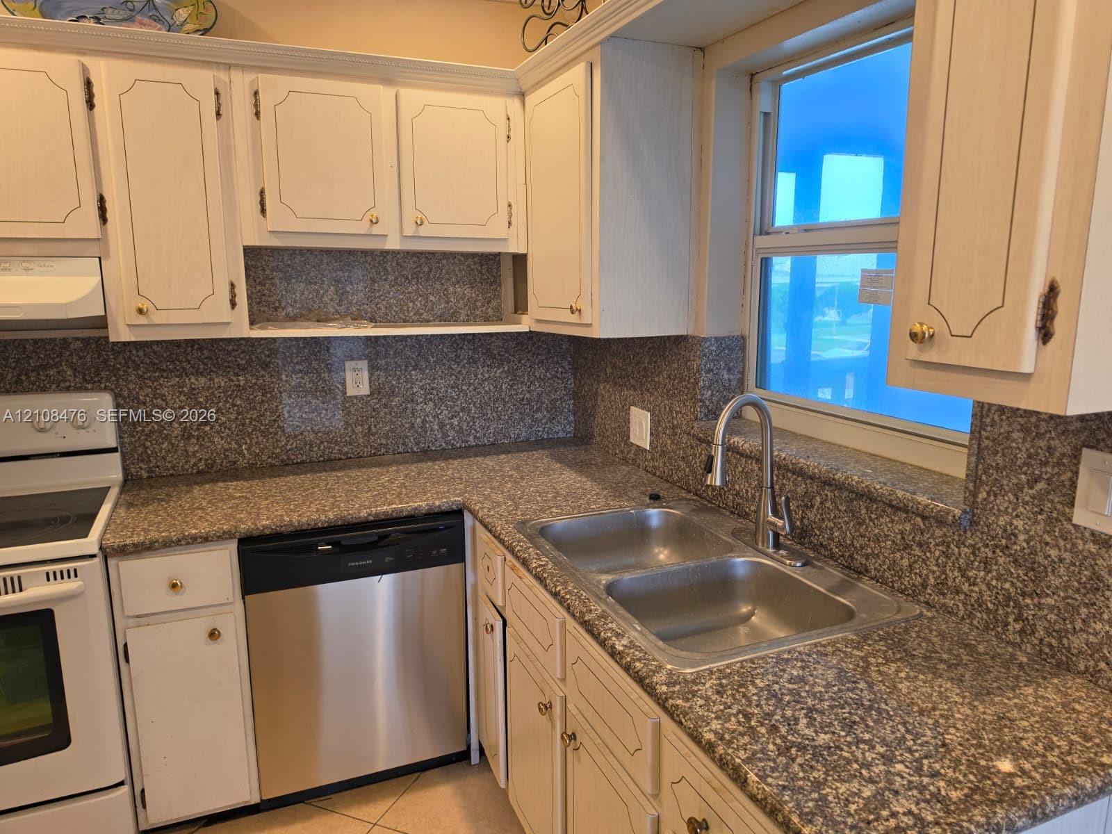 Kitchen featuring light-toned cabinetry, speckled stone-finish countertops, full-height backsplash, stainless steel dishwasher, and a dual-basin sink with a high-arch faucet Kitchen featuring light-toned cabinetry, speckled stone-finish countertops, full-height backsplash, stainless steel dishwasher, and a dual-basin sink with a high-arch faucet