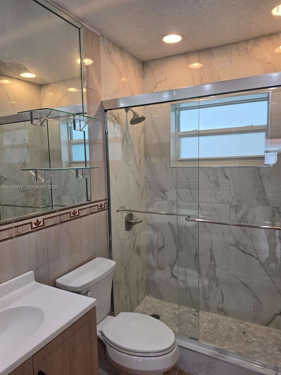 Walk-in shower featuring large-format tile surround, sliding glass door, and a pebble-stone floor finish, complemented by a light-tone vanity with a solid-surface countertop and a large wall-mounted mirror with built-in glass shelving