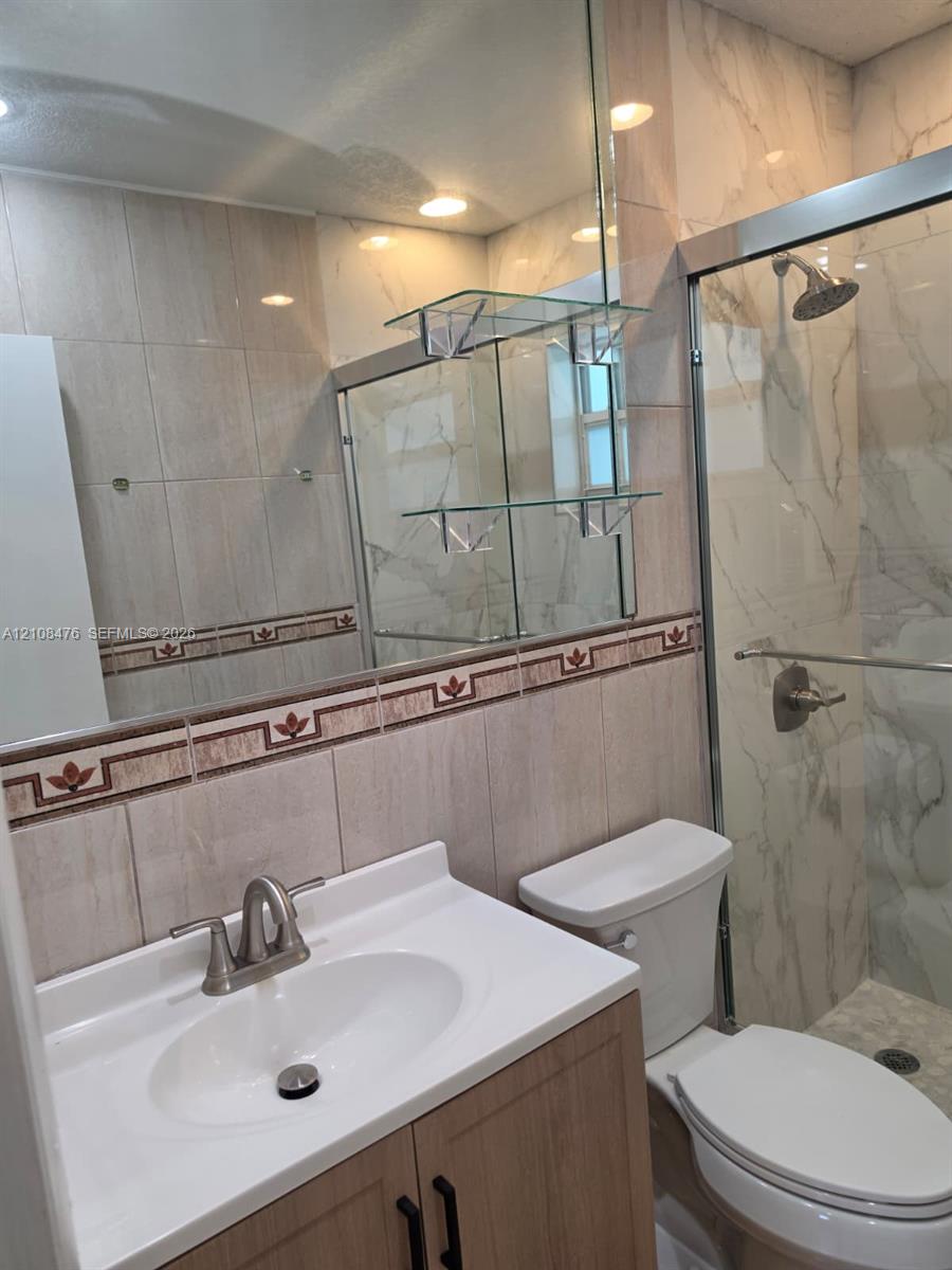 Bathroom featuring a wood-finish vanity with a white countertop, large wall-mounted mirror, glass shelving, and a shower enclosure with floor-to-ceiling decorative tile