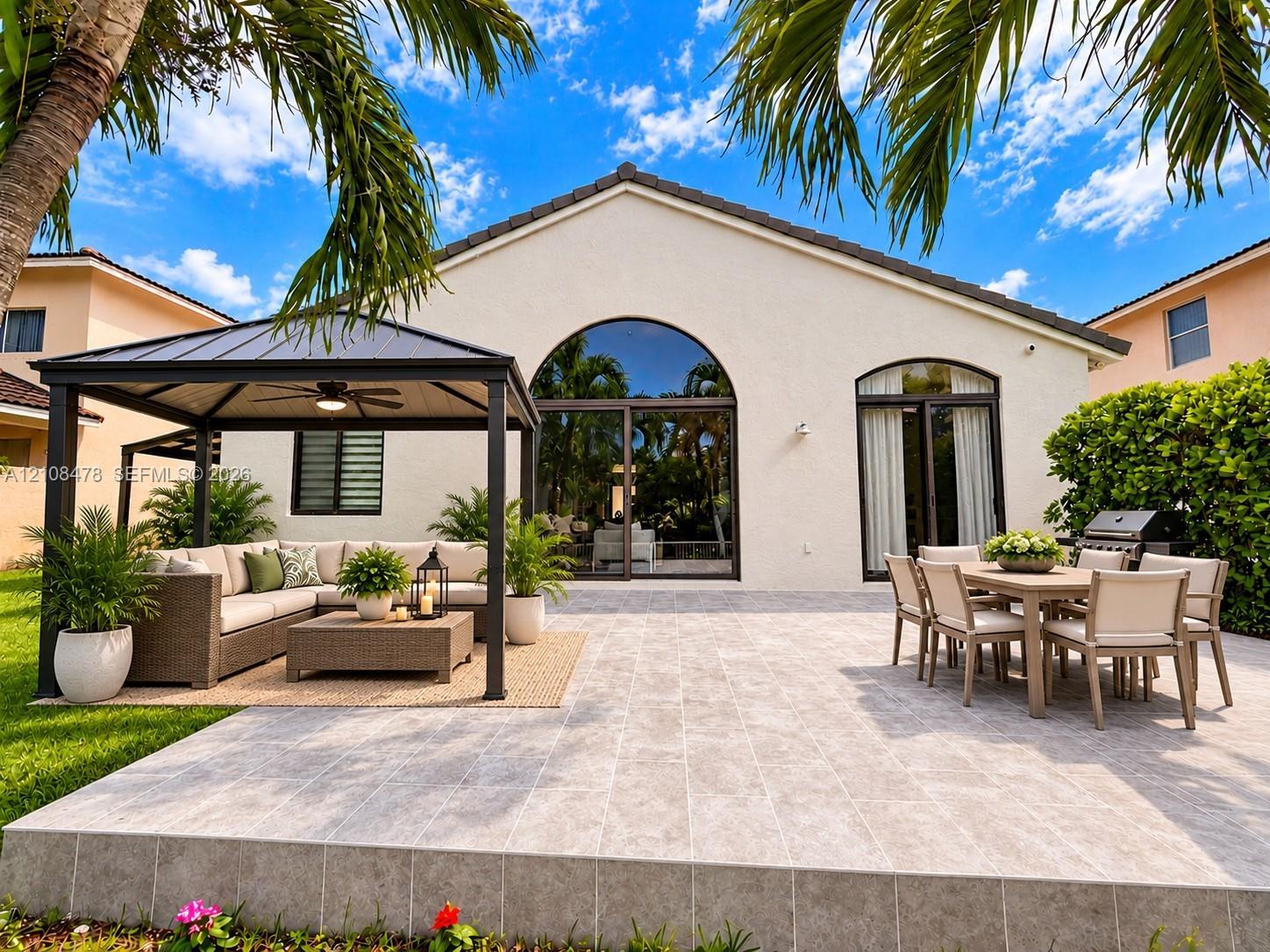 Expansive stone-paved patio featuring mature palm trees, manicured lawn, white metal perimeter fencing, and water frontage