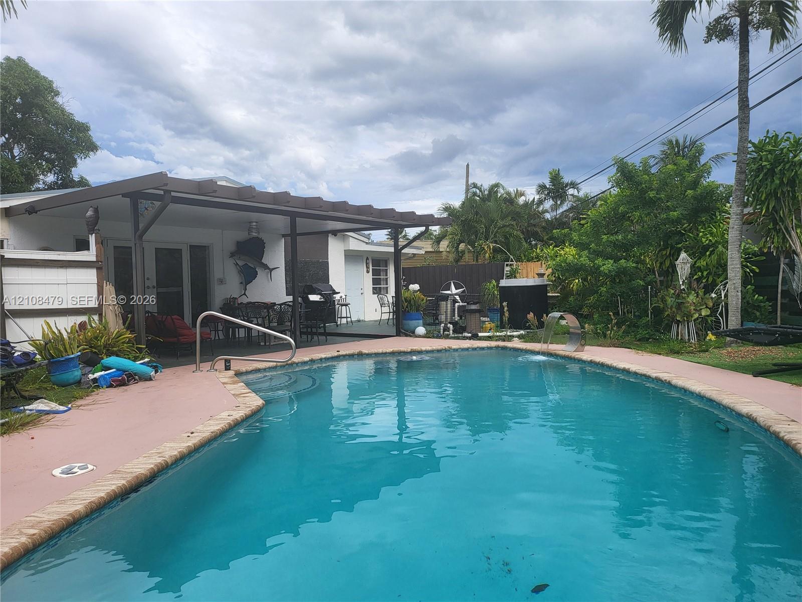 In-ground swimming pool featuring a brick-paver border and a water feature, complete with a covered patio area and mature perimeter landscaping