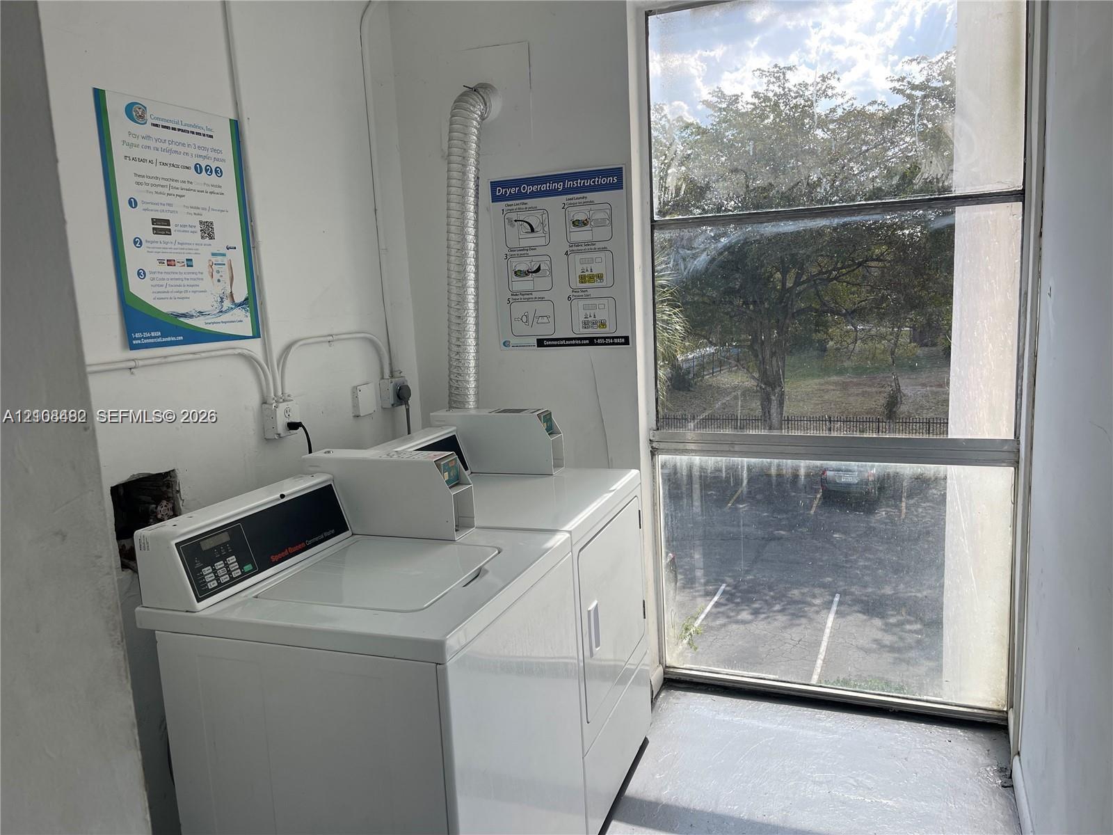 Laundry area featuring coin-operated washer and dryer units, a multi-pane fixed window, and a metal dryer vent Laundry area featuring coin-operated washer and dryer units, a multi-pane fixed window, and a metal dryer vent