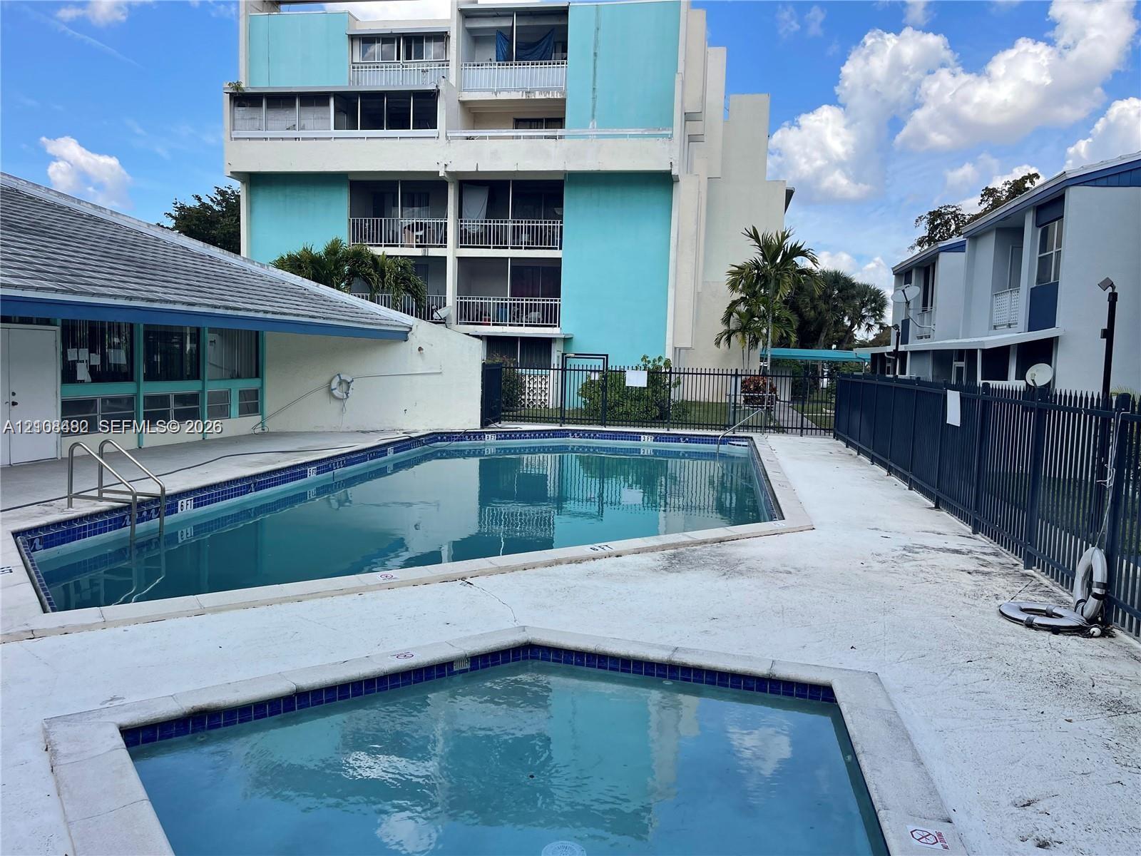 In-ground swimming pool and secondary spa featuring tile-bordered edges, concrete decking, metal perimeter fencing, and high-rise building backdrop In-ground swimming pool and secondary spa featuring tile-bordered edges, concrete decking, metal perimeter fencing, and high-rise building backdrop