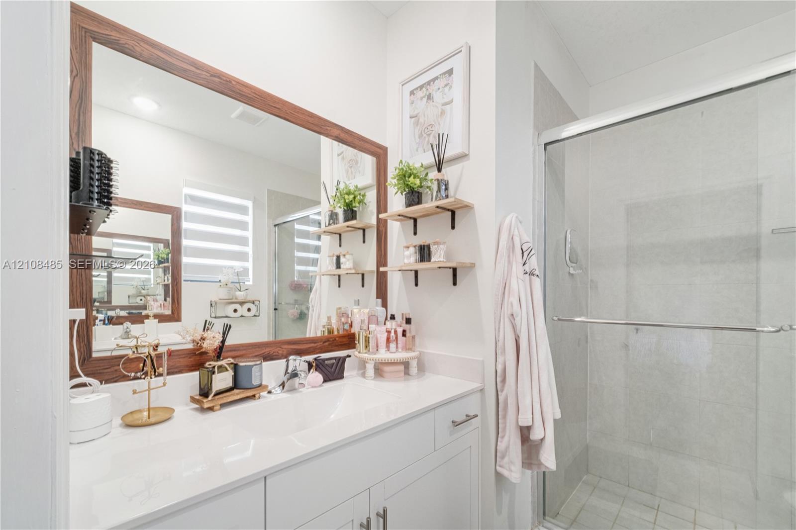 Vanity featuring a white countertop with an integrated basin, wood-frame mirror, glass-enclosed shower, and wall-mounted shelving