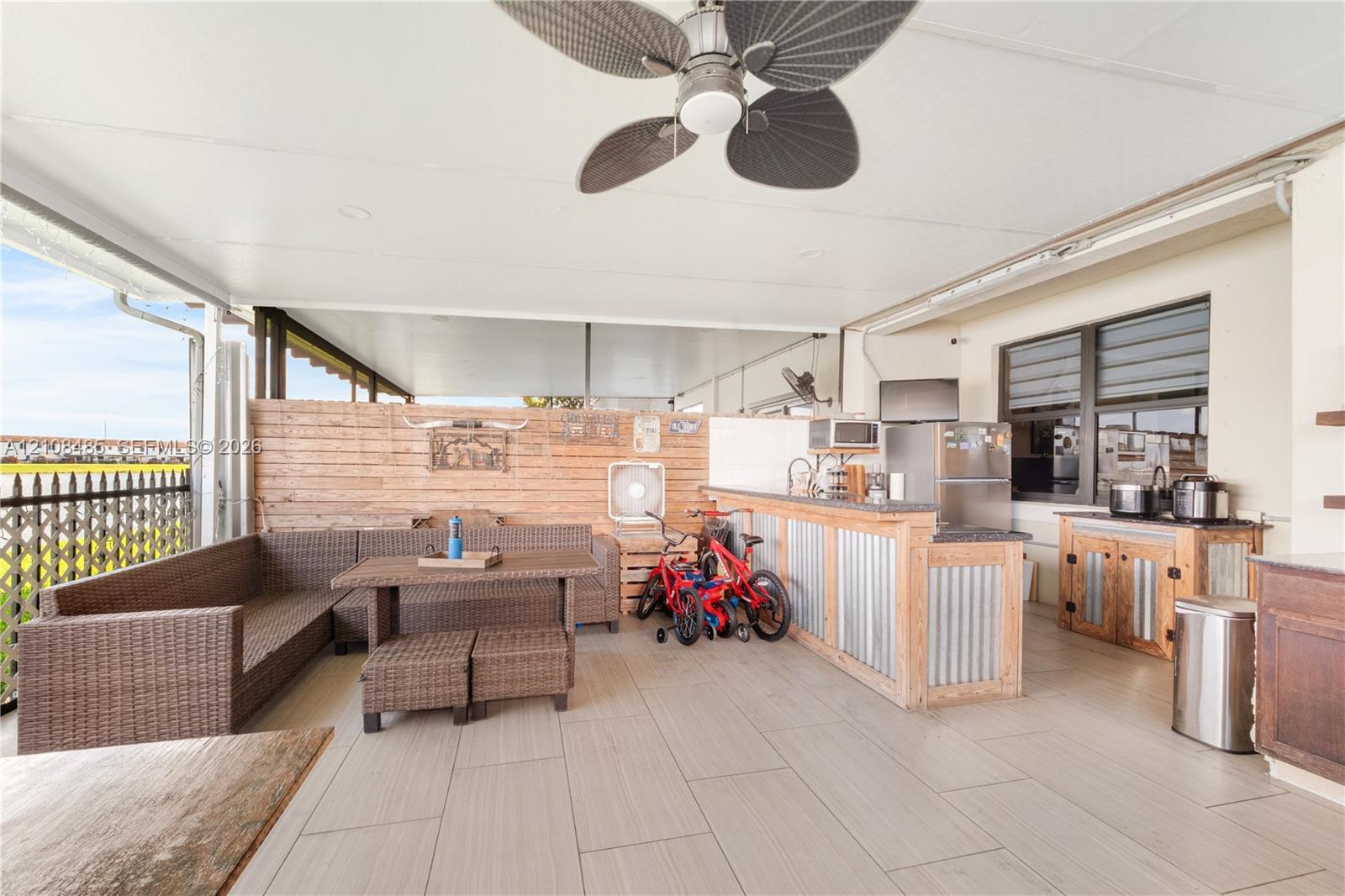 Covered outdoor patio featuring light-toned tile flooring, built-in bar with metal siding, horizontal wood-slat privacy wall, ceiling fan, and louvered windows