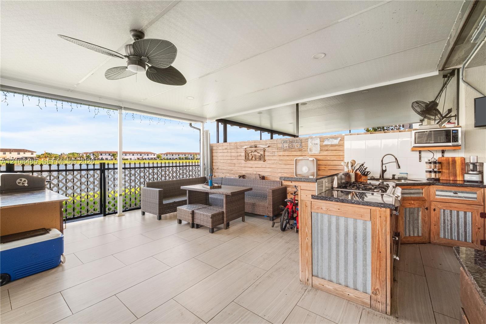 Covered patio featuring large-format tile flooring, a ceiling fan, and a built-in outdoor kitchen with stone-finish countertops, wood cabinetry with metal paneling, and a high-arch faucet