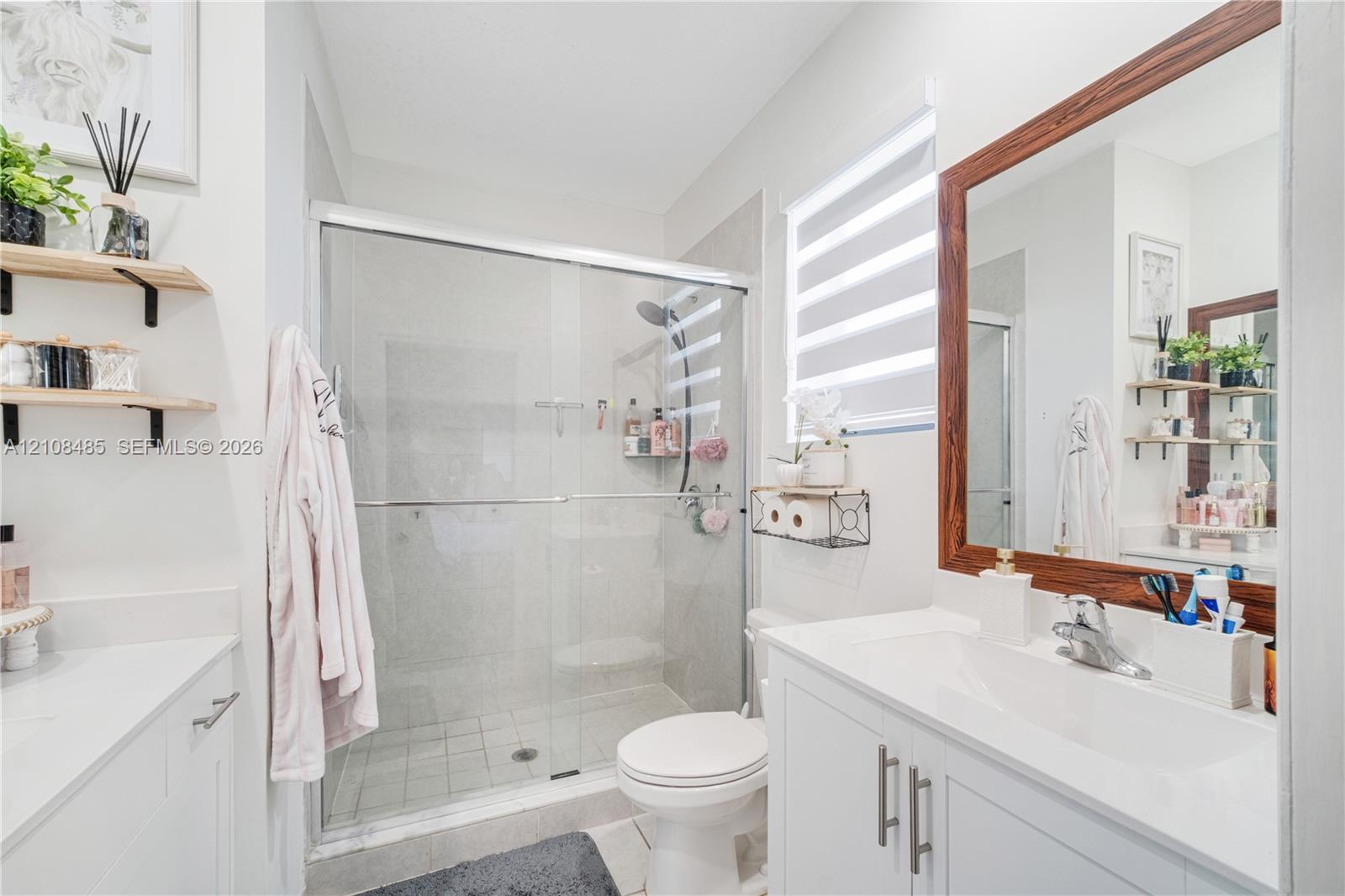 Bathroom featuring a vanity with a white countertop and rectangular undermount sink, a large wood-framed mirror, walk-in shower with glass enclosure, and wall-mounted shelving