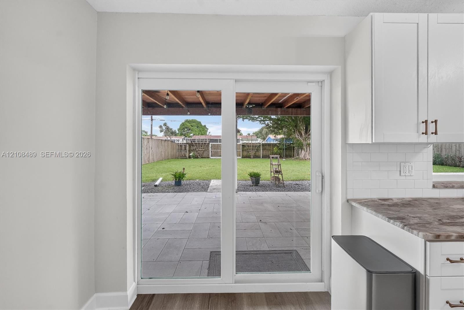 Sliding glass door opening to a concrete patio, covered pergola, fenced backyard with turf, and white shaker-style cabinetry with subway tile backsplash