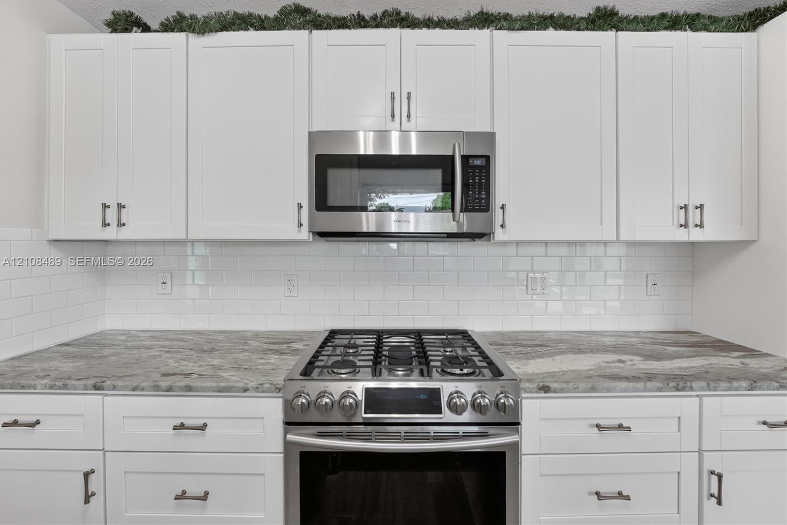 White shaker-style cabinetry with metallic hardware, stone-finish countertops, subway tile backsplash, and stainless steel gas range and built-in microwave