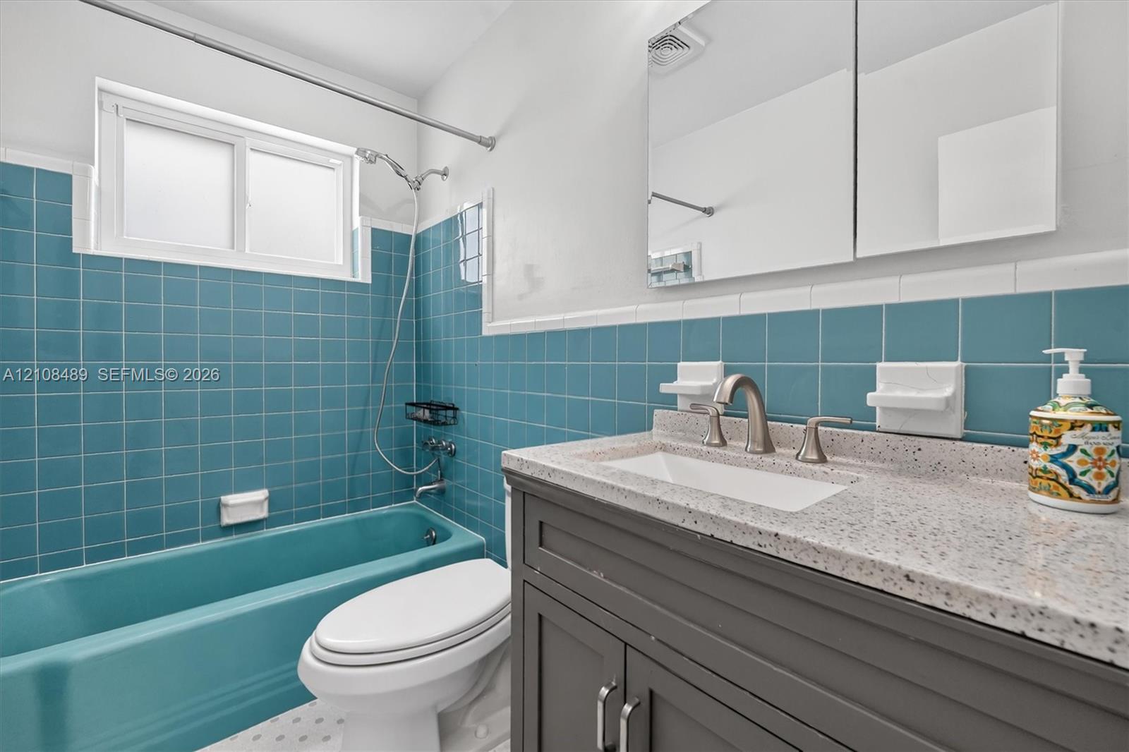 Bathroom featuring a vanity with speckled countertop, integrated sink, and charcoal cabinetry, complemented by teal wall tiling, a matching teal soaking tub, and a wall-mounted mirrored medicine cabinet