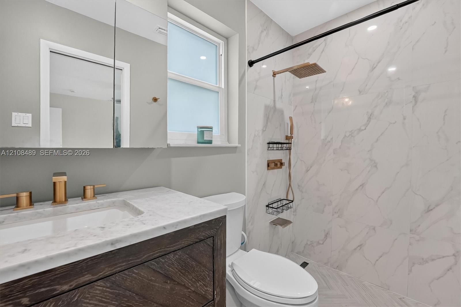Bathroom featuring a vanity with wood-finish cabinetry, a light-toned stone countertop, brass-finish fixtures, a shower enclosure with large-format stone-patterned wall tile, and herringbone-patterned floor tile