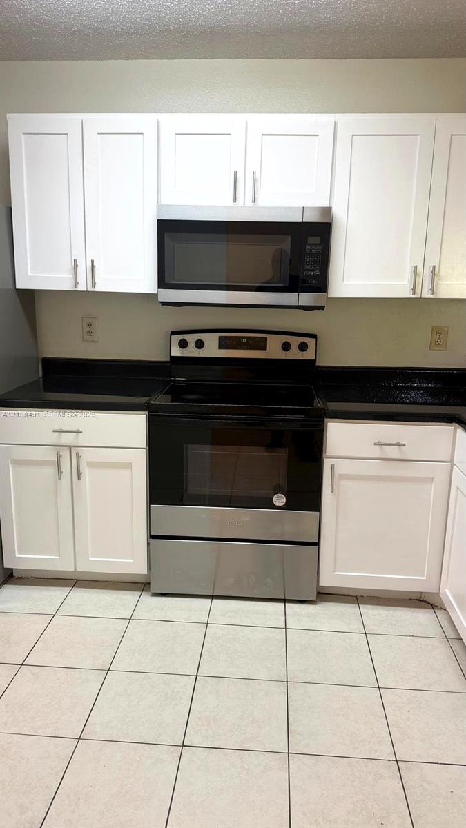 Kitchen featuring white shaker-style cabinetry, dark stone-finish countertops, stainless steel appliances, and large-format ceramic tile flooring Kitchen featuring white shaker-style cabinetry, dark stone-finish countertops, stainless steel appliances, and large-format ceramic tile flooring