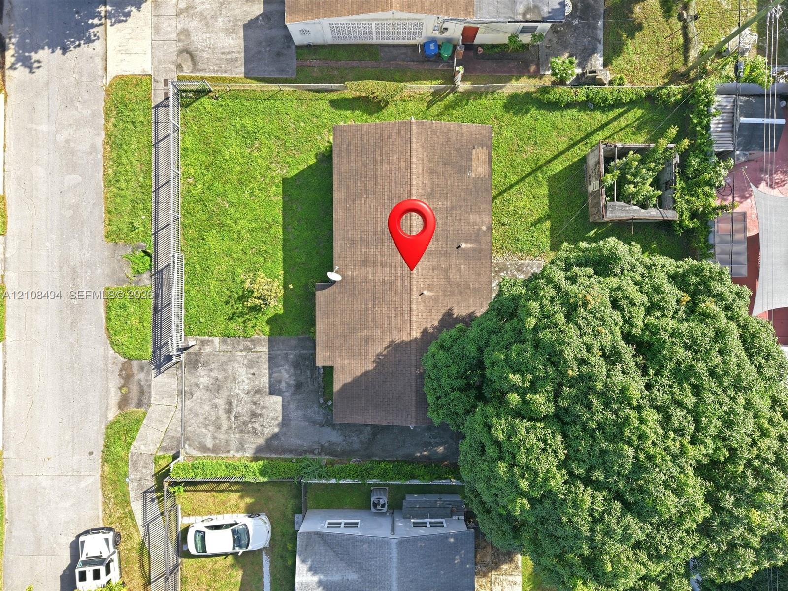 Aerial perspective of property lot featuring an expansive lawn, concrete driveway, perimeter chain-link fencing, and mature trees