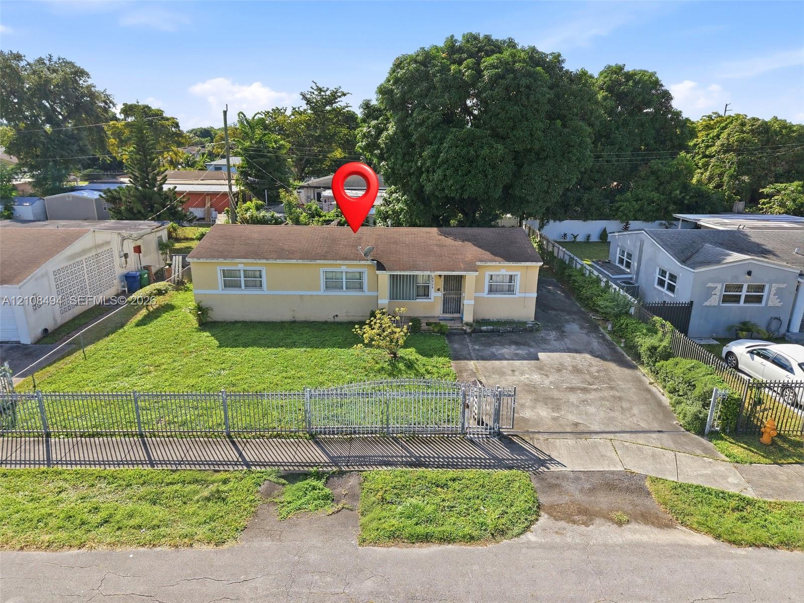 Single-story residence featuring a low-slope roof, stucco exterior, paved driveway, metal perimeter fencing, and mature trees Single-story residence featuring a low-slope roof, stucco exterior, paved driveway, metal perimeter fencing, and mature trees