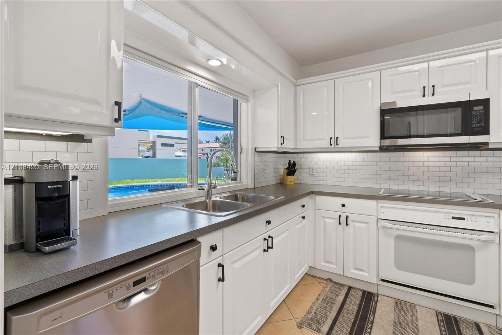 Kitchen featuring white shaker-style cabinetry, solid-surface countertops, a subway tile backsplash, and a stainless steel dishwasher and microwave