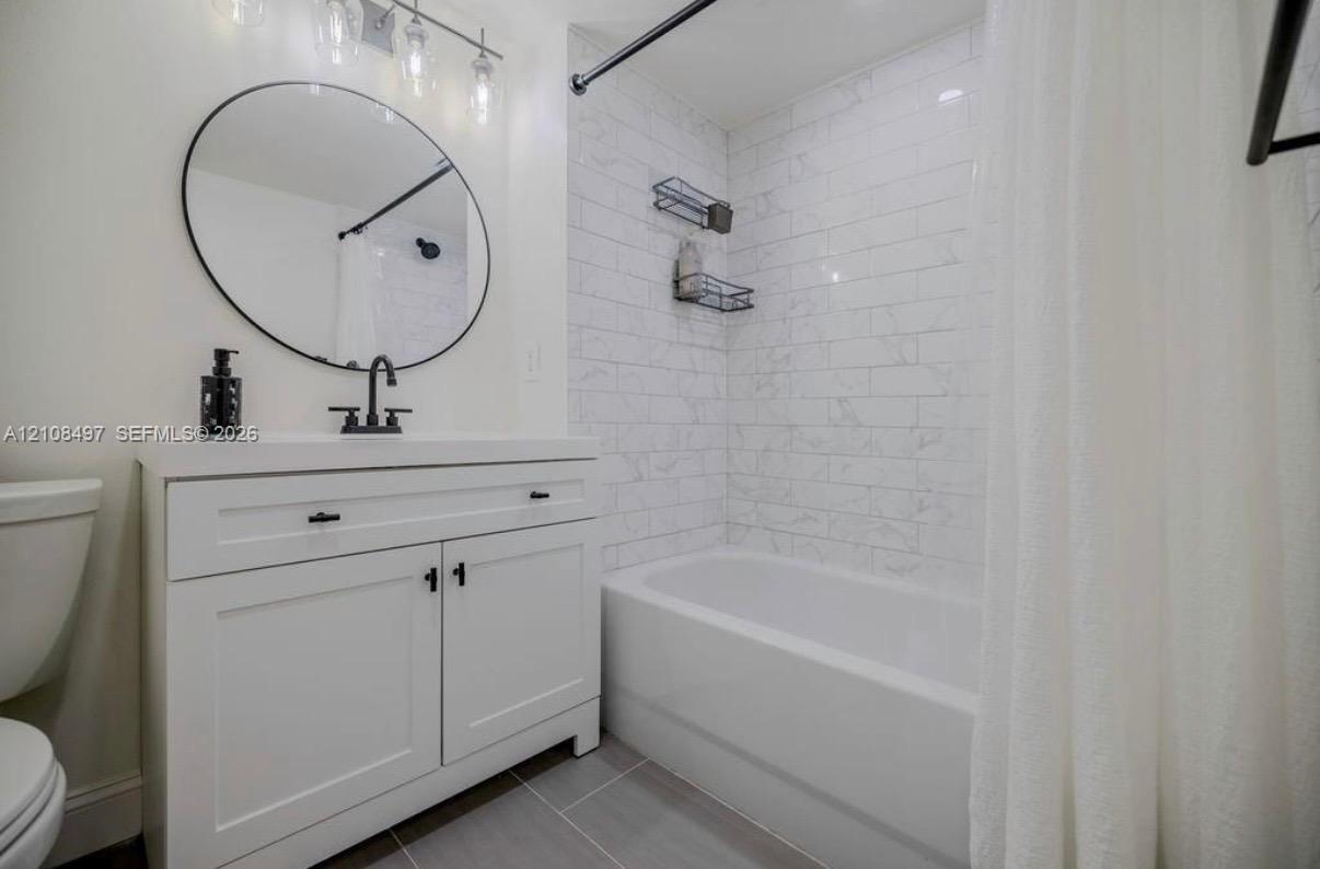 White vanity with shaker-style cabinetry, matte black faucet, round wall-mounted mirror, light-toned subway tile shower surround, and grey tile flooring