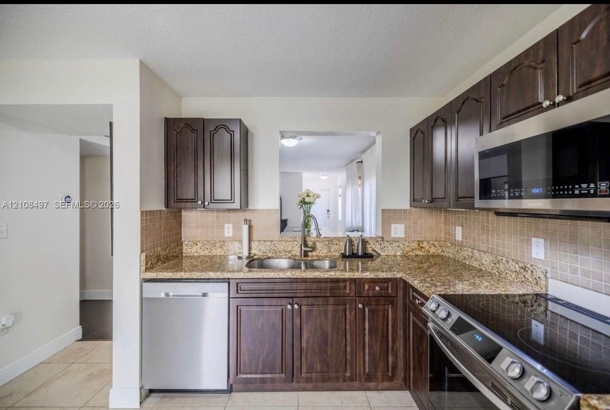Kitchen featuring dark-finish wood cabinetry, stone-surface countertops, square tile backsplash, and stainless steel appliances including a built-in microwave and dishwasher